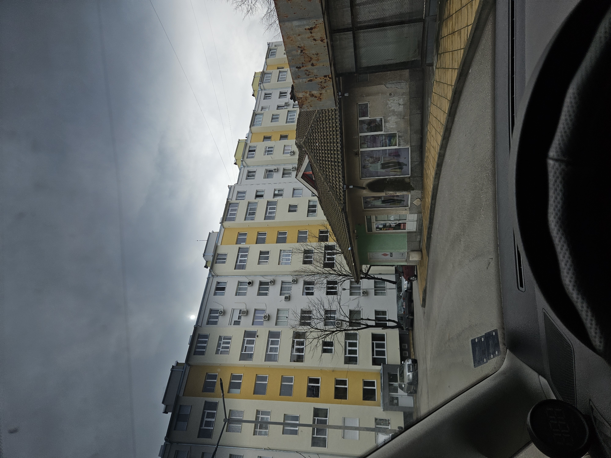 The image, taken from inside a vehicle, shows a multi-story apartment building in Haskovo, Bulgaria. The building has a yellow and white facade and numerous windows with air conditioning units attached. Bare tree branches are visible in front of the building. Adjacent to the apartment building is a lower, single-story structure with commercial storefronts. The sky is overcast, with gray clouds obscuring the sun. Part of the vehicle's interior, including the steering wheel, is visible in the foreground. Several cars are parked on the street in front of the building. The environment appears urban and possibly residential.