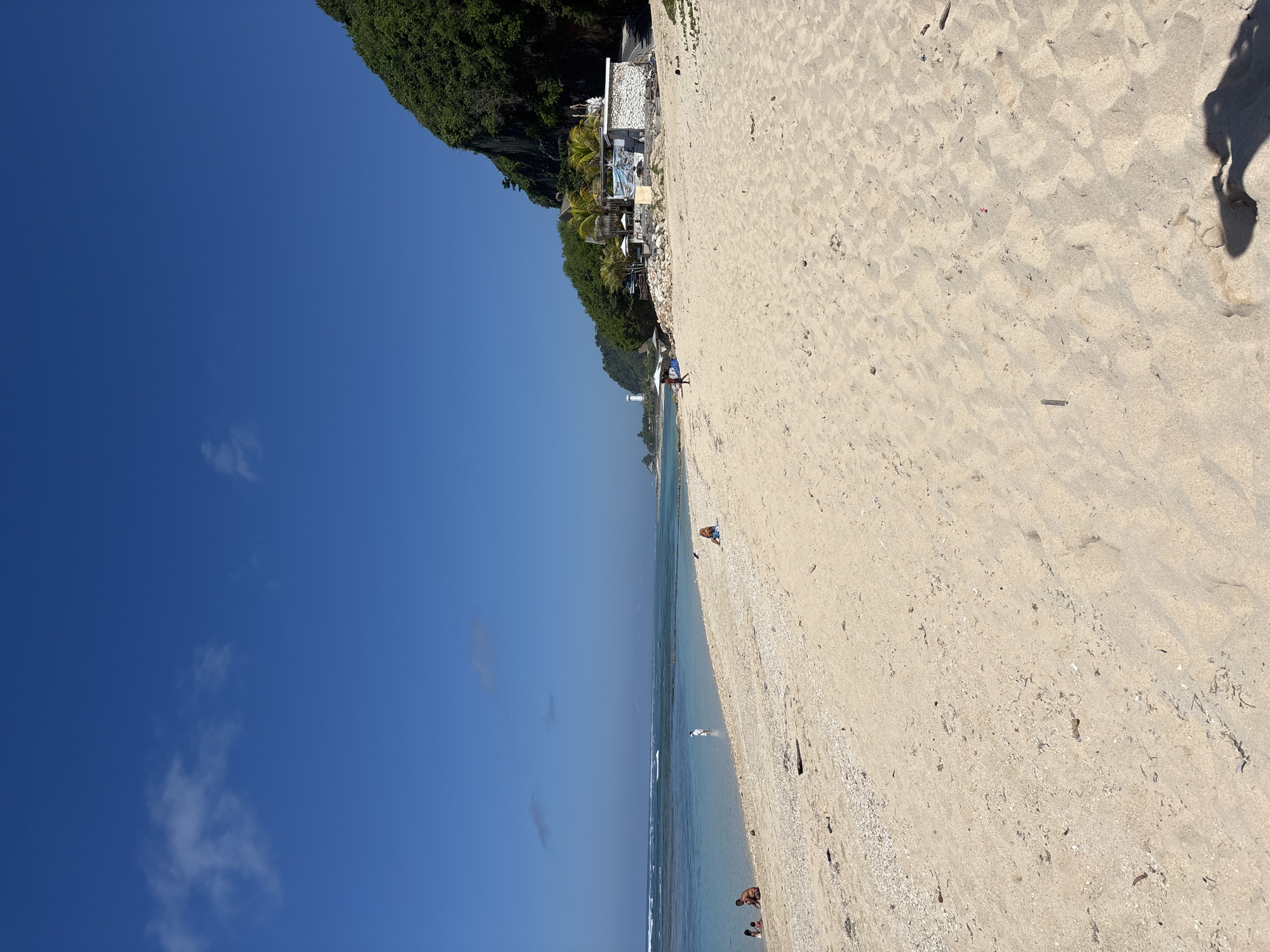 The image shows a beach scene in Kutuh, Indonesia. The beach is sandy with a high angle view showing the beach from top to bottom. There are people on the beach and in the water. There is a shadow of a person in the top right corner, implying the photographer is standing to the right. The sky is blue with some clouds. A green cliff is to the left, which appears to have a building.