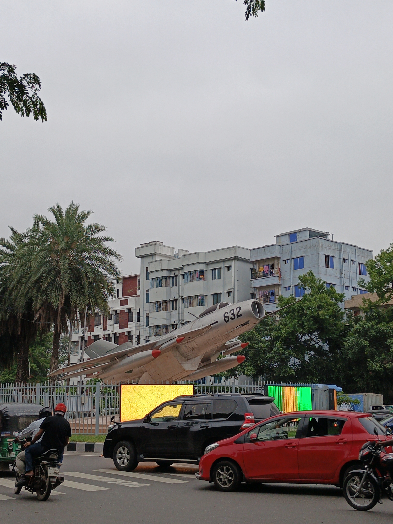 The image presents a bustling urban street scene in Dhaka, Bangladesh, under an overcast sky. A prominent feature on the left is a light-colored fighter jet, possibly a MiG-15 or MiG-17, displayed as a monument on an elevated structure. The jet has the number "632" painted near its nose intake and distinctive red tips on its wings.
Behind the jet and along the street, several multi-story buildings rise, varying in shades of light blue and white with red accents. Clothes are visible drying on some balconies, suggesting residential occupation. Abundant green foliage, including a tall palm tree on the left and other leafy trees, adds a tropical touch to the cityscape.
The foreground and mid-ground of the street are active with vehicles. A red hatchback car is centrally positioned, while a black SUV is partially visible behind it, seemingly next to a large, bright yellow illuminated display or billboard. On the left, a motorcycle with two riders—one wearing a red helmet—is seen near a zebra crossing, with a green auto-rickshaw partially visible behind it. Another motorcycle is on the far right.
The scene captures a typical, busy day in an urban environment, with the fighter jet serving as a distinctive landmark amidst the flow of daily city life.