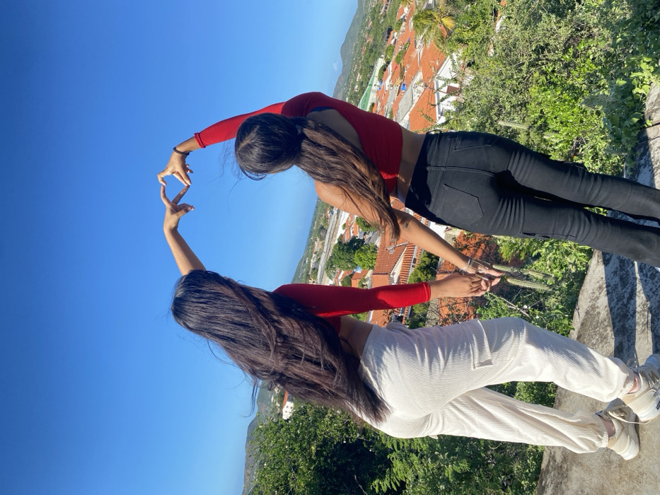 Two women stand closely together, facing away from the camera, wearing matching red long-sleeve crop tops. The woman on the right wears black jeans, while the other has on cream-colored corduroy pants. They extend their arms upwards, hands meeting to form a heart shape against a bright blue sky. In the background, a cityscape with orange-tiled roofs and lush green trees is visible, suggesting a high vantage point.
