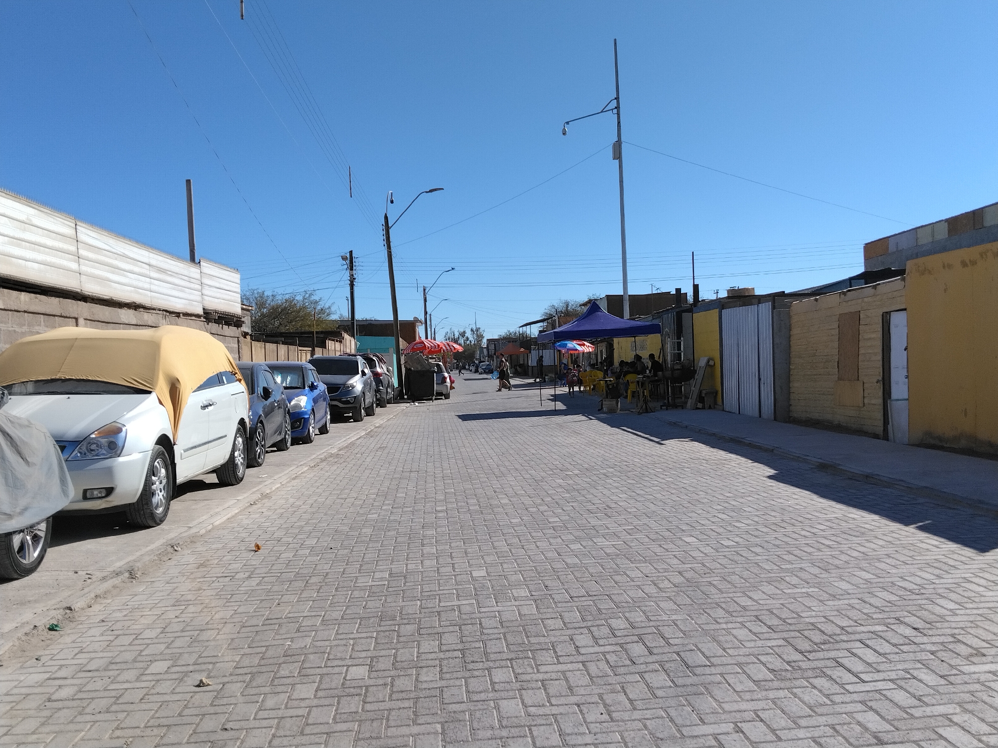This image depicts a street scene in Pozo Almonte, Chile. The street is paved with interlocking bricks, and on the left side, several cars are parked along the sidewalk. One car is partially covered with a yellow tarp. The buildings lining the street appear to be simple structures, some with corrugated metal siding and others with yellow or beige painted facades.
In the distance, there are more parked cars and a few people walking along the sidewalk. On the right side of the street, there are some market stalls with blue and red umbrellas providing shade. Several people are gathered at these stalls, suggesting a commercial or social gathering. The sky is clear and blue, indicating a sunny day. The presence of utility poles and wires suggests an urban or semi-urban environment. There is no visible text in the image. The overall impression is that of a typical daytime scene in a small town or city.