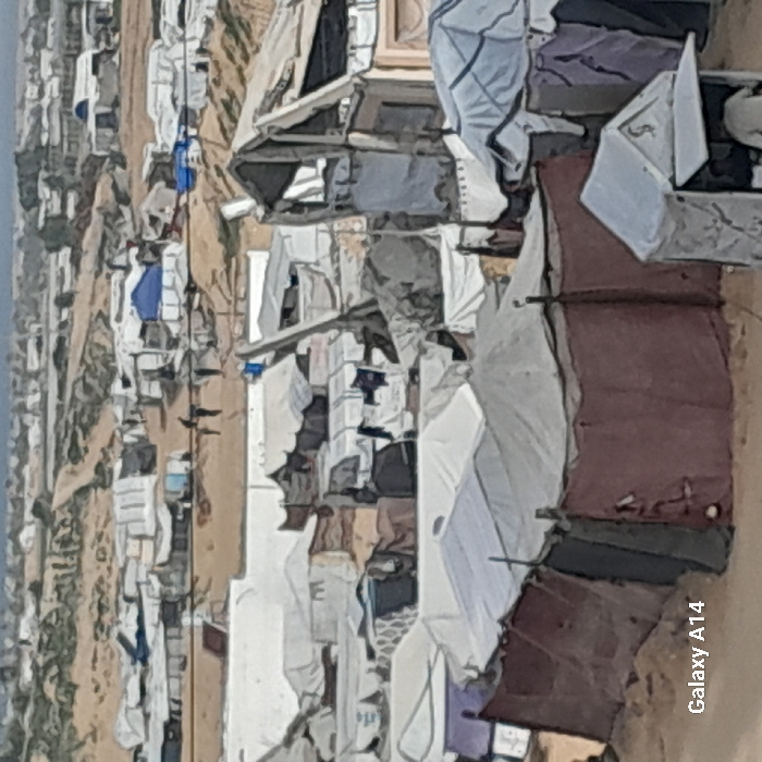 The image depicts a densely populated refugee camp. Makeshift shelters, constructed from tarps, cloth, and scavenged materials, are clustered closely together on barren land. Rows of tents extend into the distance, suggesting a large-scale displacement situation. Clothing is hung out to dry, and miscellaneous belongings are piled up around the shelters. The overall impression is one of poverty, overcrowding, and hardship. The text "Galaxy A14" appears on the lower right, likely indicating the device used to take the photo.
