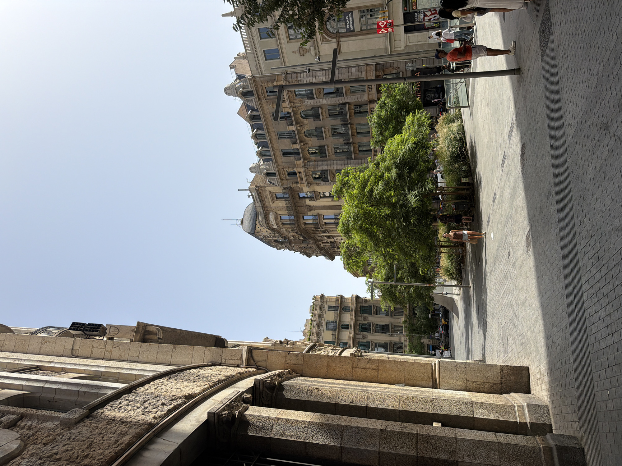 The image depicts a sunlit urban scene in Barcelona, Spain, likely on a sidewalk. On the left, a portion of a building facade with large, fluted stone columns and detailed architectural carvings is visible. In the center and right of the frame, several multi-story buildings with ornate facades, balconies, and mansard roofs line a street. Lush green trees and bushes are strategically planted in a public area, creating a contrast with the urban architecture. Several individuals are scattered throughout the scene, some walking and others standing. A woman in a light-colored top and shorts stands in the foreground on the right, holding a bag. Further down the street, more people are visible, including a man in an orange t-shirt and white shorts. The ground is paved with cobblestones and features metal gratings. A sign with the Barcelona Metro logo is visible on the right side of the image, confirming the location. The sky is clear and bright blue, indicating fair weather.