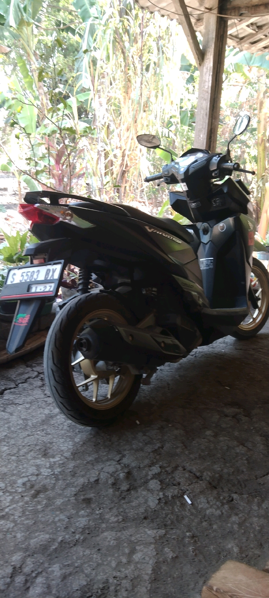 A black motorcycle with gold rims is parked on a cracked concrete surface. The motorcycle is facing to the right. It has a license plate that reads "S 5583 BX" and the date "11-27". The motorcycle is under a wooden overhang, with lush green foliage visible in the background, suggesting an outdoor setting in a tropical or sub-tropical climate. The overall impression is of a scene in a residential or rural area, possibly a yard or alleyway, within the city of Wadang, Indonesia.
