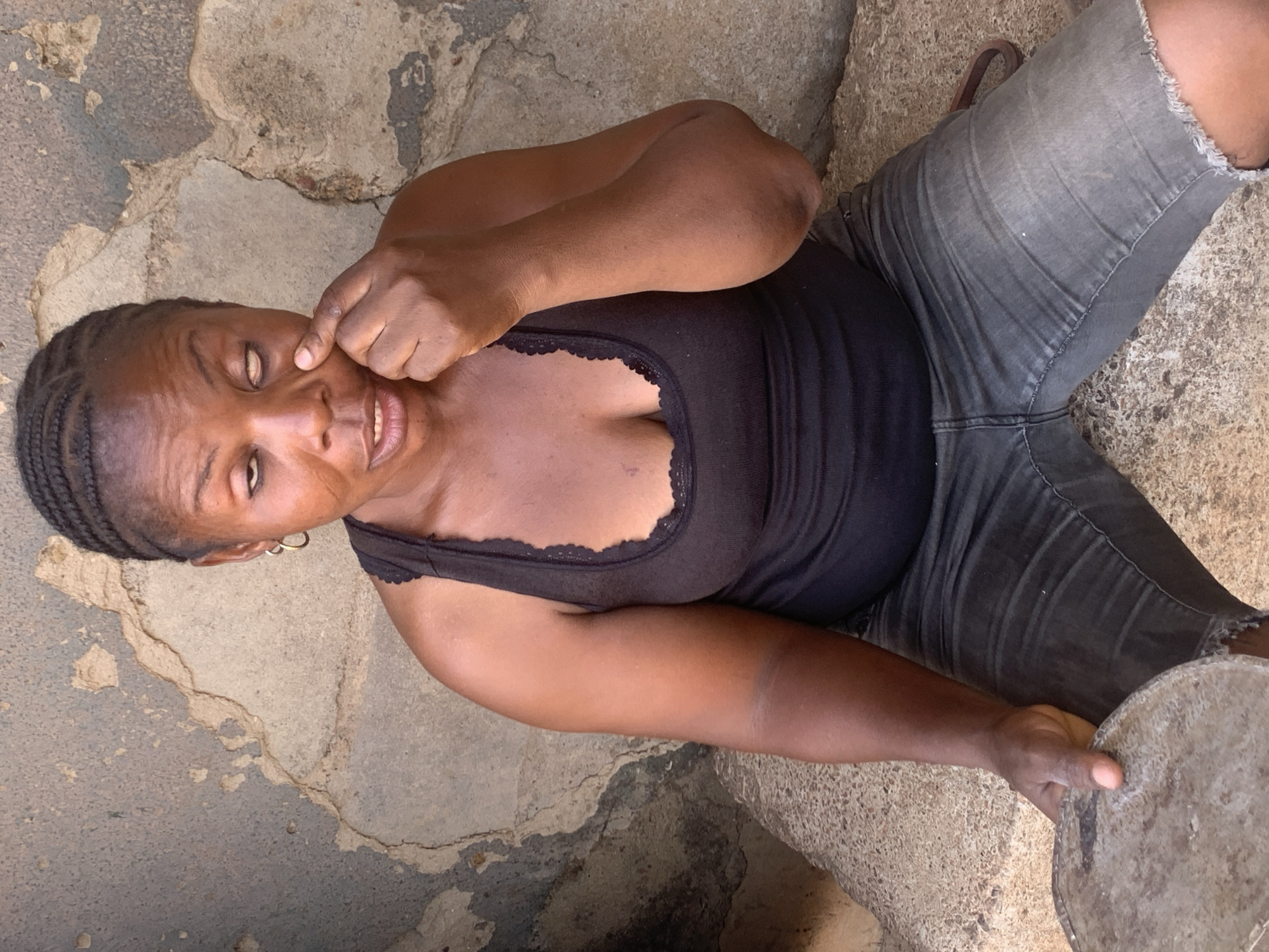 A woman is sitting on a cracked stone or concrete surface, possibly outdoors. She wears a black tank top with lace trim and grey denim shorts. The woman is touching her face near her eye with her right hand. In her left hand, she holds a circular, grey object. Her hair is braided. The location is not specified in the original request.