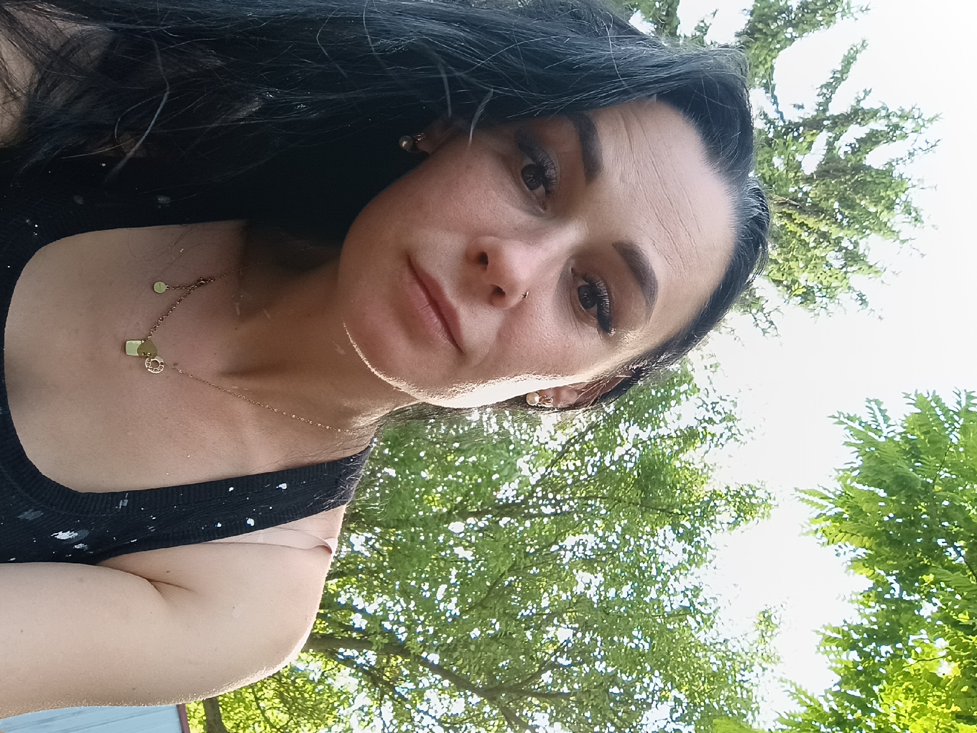 A woman with long dark hair and noticeable makeup, including dramatic eyelashes and defined eyebrows, poses for a selfie outdoors. She is wearing a black tank top with white splotches and a gold necklace with small circular and rectangular pendants. A subtle nose ring is visible. Her left earlobe has a small, pearl-like earring. The background consists of bright green foliage from trees and a light-colored sky, suggesting a sunny day. The overall setting appears to be a natural, outdoor environment, possibly a yard or park, in or near Kliushnykivka, Ukraine. The image is taken from a slightly high angle, looking down at the woman's face.