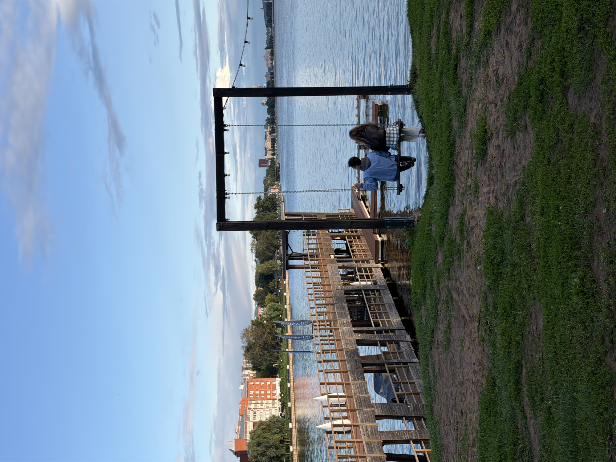 Two individuals are seated on a swing overlooking a body of water. The swing set, constructed of dark wood, has a horizontal bar with hanging lights. The individuals are positioned with their backs to the camera, one wearing a blue jacket and the other a dark jacket over a plaid skirt. They are on a sandy area with grass in the foreground. In the background, a wooden pier with railings extends into the water, lined with white vertical markers and two blue flags. Across the water, buildings and trees are visible on the opposite shore, consistent with the shoreline of Votkinsk, Russia. The sky above is blue with scattered clouds.
