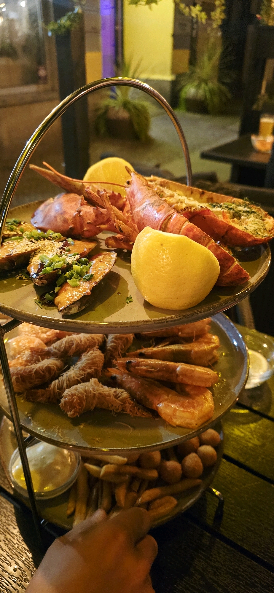 A three-tiered metal serving stand holds a seafood platter, fries, and a small bowl of sauce on a dark wooden table. The top tier features a whole lobster, two lemon wedges, and several pieces of what appear to be glazed sweet potato topped with sesame seeds and chopped scallions. The middle tier displays a generous portion of cooked shrimp and some crispy, shredded pastry-wrapped items. The bottom tier is laden with french fries and small, round fried items, possibly arancini or hushpuppies, and a small bowl of yellow sauce. The scene is dimly lit, suggesting an evening setting, possibly a restaurant with ambient lighting, indicated by a blurred background with yellow and purple hues from external lights. A person's hand is visible in the lower right corner, reaching towards the bottom tier.
