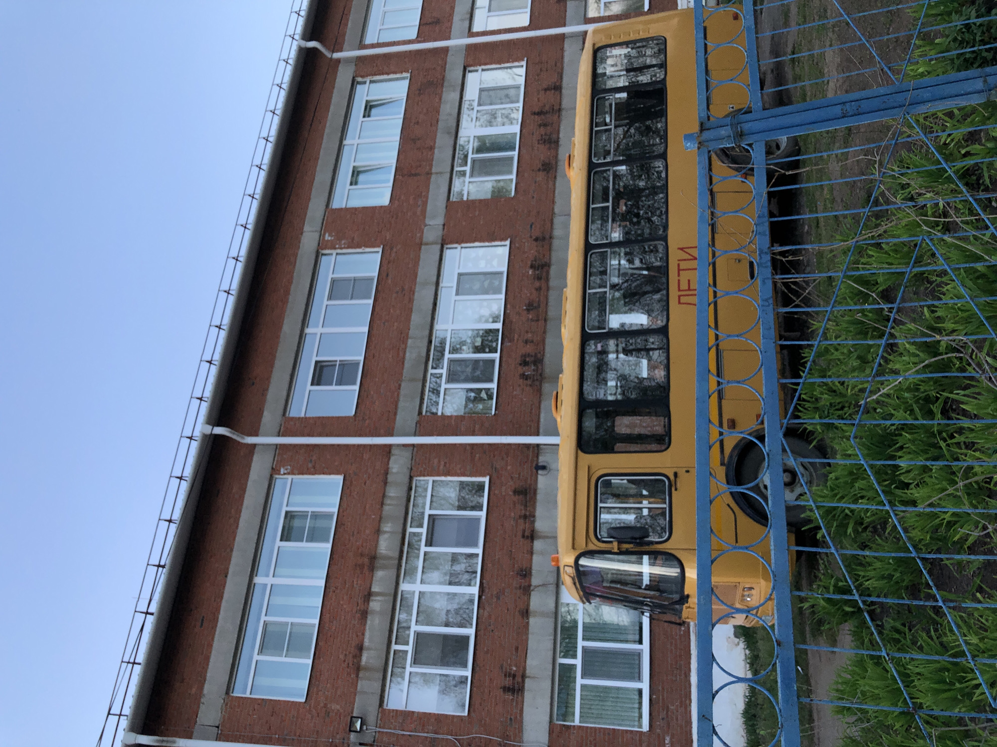 A yellow school bus with the word "ДЕТИ" (children) written on its side is parked adjacent to a red brick building with multiple windows. A blue metal fence partially obscures the bus. The scene is set outdoors on a clear day under a blue sky. The location is Noviy, Russia.
