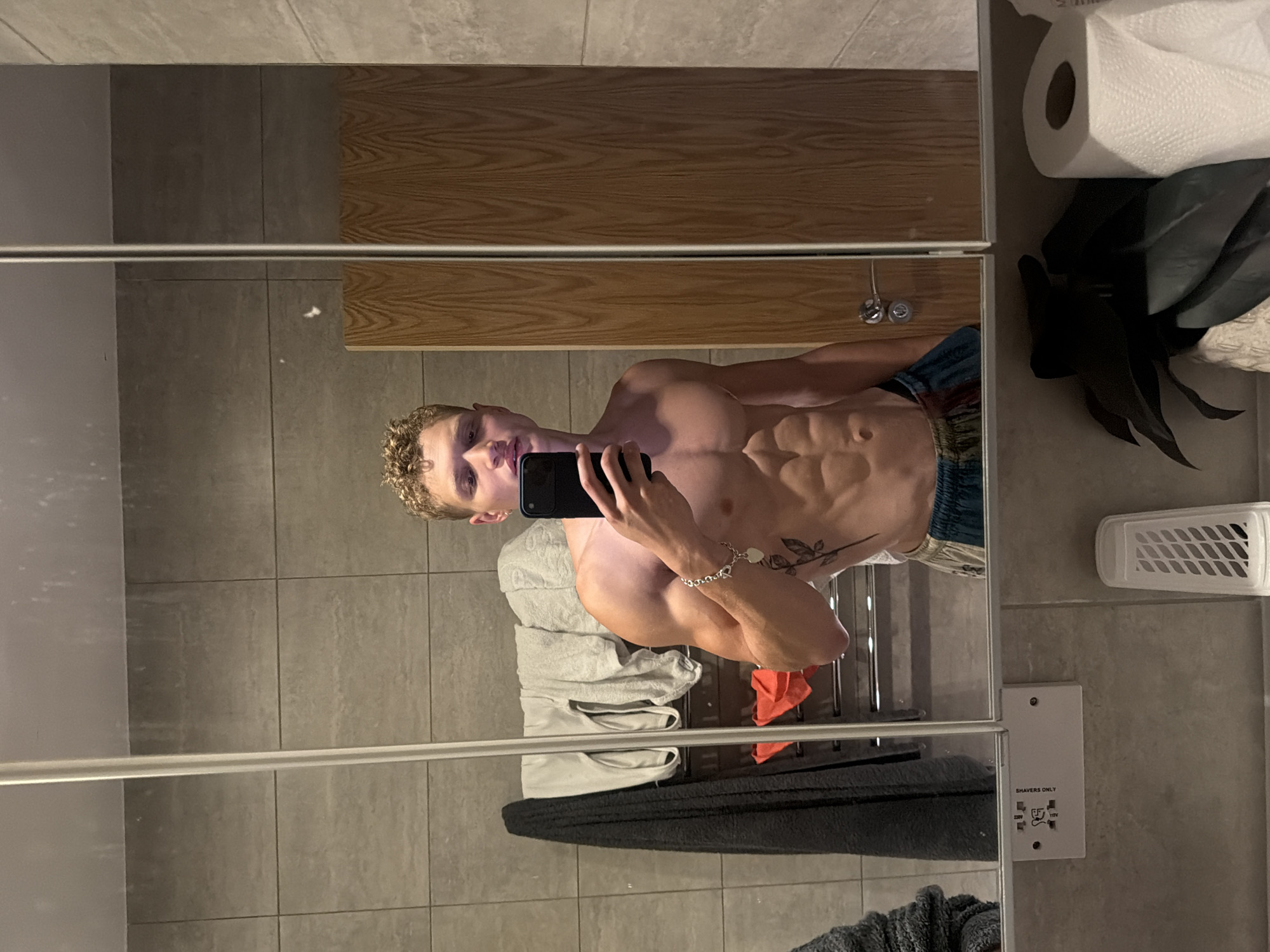 A light-skinned male, appearing to be in his twenties, is depicted from the waist up in a mirror selfie taken within a bathroom. He possesses curly blonde hair and a lean, muscular physique with visible abdominal definition. He is shirtless, wearing multi-colored patterned shorts. A silver-toned bracelet with heart-shaped charms is present on his left wrist, and a dark tattoo resembling a plant stem with leaves is visible on his left torso. He holds a black smartphone in his right hand to capture the image, with his gaze directed towards the phone.
The bathroom features light grey square wall tiles and a multi-panel mirrored cabinet above a grey countertop. Reflected in the left-hand mirror panel is a chrome heated towel rail holding several towels, including dark grey, light grey, and a small orange towel. Below the left mirror panel, a white wall-mounted shaver socket is visible, marked "SHAVERS ONLY" with both "230V" and "115V" inputs, characteristic of electrical fittings in the United Kingdom. Reflected in the right-hand mirror panel is a wooden door or cabinet interior with a circular silver handle. On the countertop, a roll of white toilet paper stands vertically to the right, adjacent to a small dark artificial plant in a white textured pot. A white slatted container is partially visible on the far left of the countertop. The scene is consistent with an interior bathroom in London, United Kingdom.