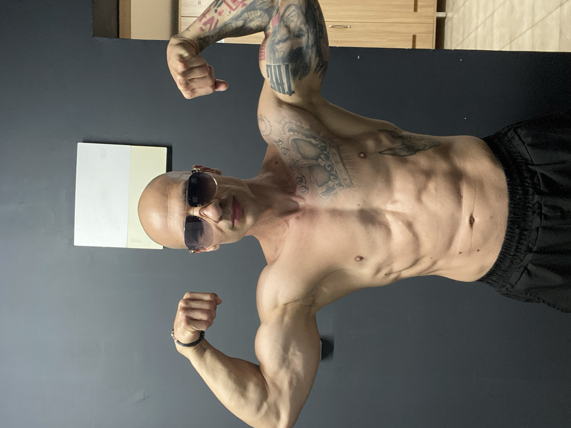 A bald, fit man with sunglasses and a crown tattoo on his chest poses with his arms flexed, displaying his biceps. He is wearing black shorts. The background is a dark gray wall with a white and beige rectangular sign, and on the right, wooden cabinets and lockers are visible. The scene appears to be inside a gym or locker room. The image's metadata indicates it was taken in Pleven, Bulgaria.