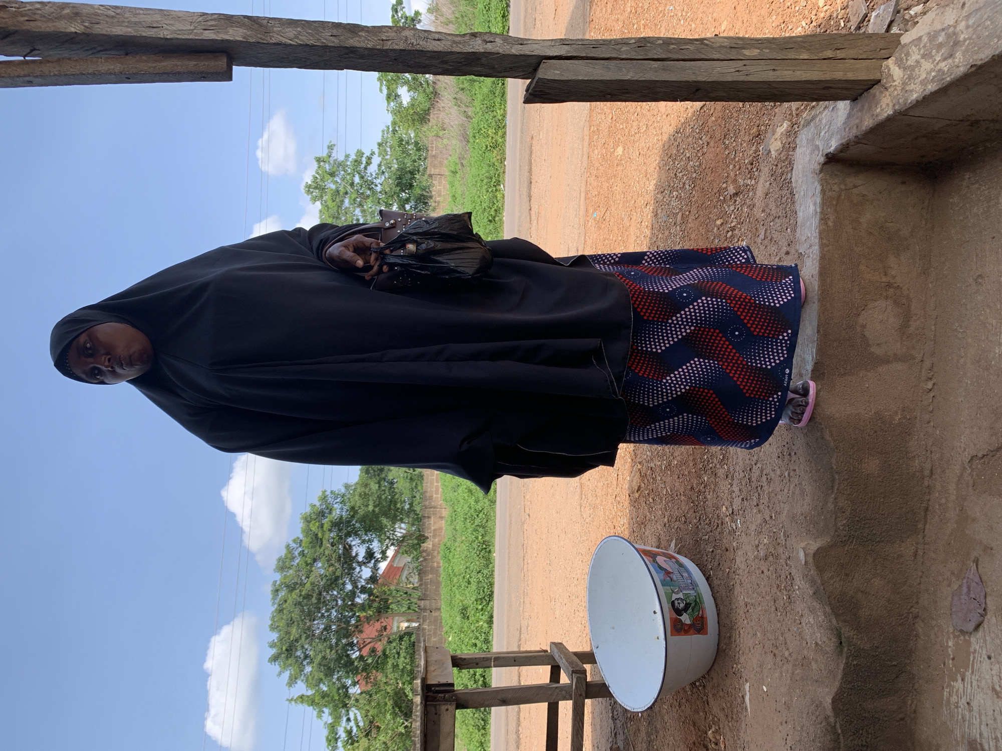 A woman stands outdoors wearing a black hijab and a patterned skirt. She holds a black bag in her right hand. In front of her, to her left, is a white oval basin with colorful images on its side. The ground is covered in reddish-brown dirt. Behind the woman, a road is visible, bordered by green vegetation and trees. In the background, a building with a red roof can be seen. The scene appears to be in Gbeleko, Nigeria, based on the context provided. The sky is blue with scattered white clouds. The woman is standing on a raised concrete platform. A wooden post is visible to the right of the frame.