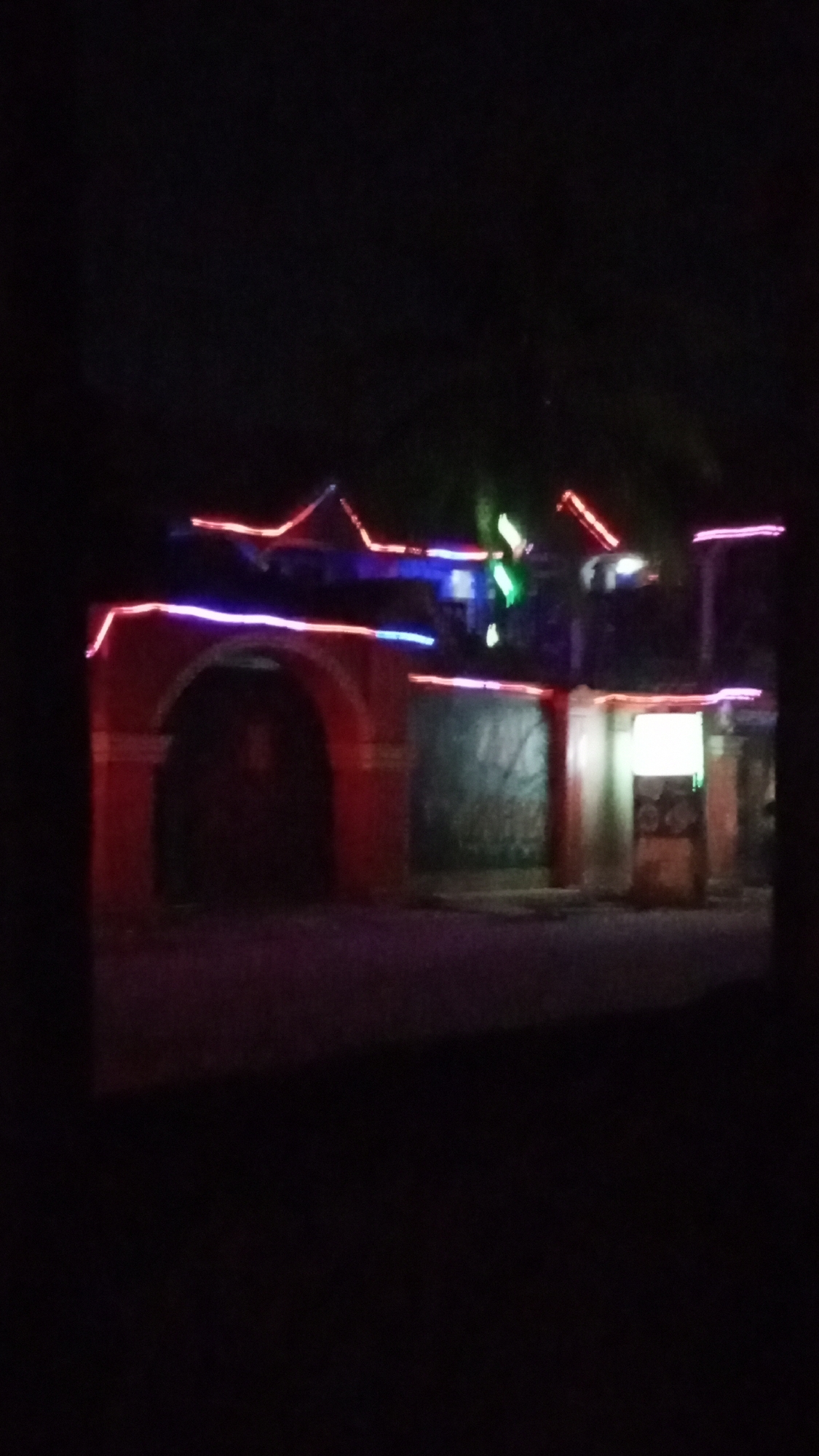 The media file captures a nighttime scene, likely in Sabon Tasha, Nigeria, featuring a building or an entrance structure illuminated by colorful lights. The central subject is a reddish-brown structure with a large, dark arched entrance in the center-left. This structure, along with an adjacent part of the building to its right, is outlined with strings of red and blue neon-like lights. Red lights trace the gabled rooflines, while blue lights run horizontally along the eaves and above the archway.
On the far right, another section of the building or a small kiosk is visible, featuring a bright, square-shaped light that appears to be part of a sign or display. Some green lights are also faintly visible, possibly from another decorative element or sign. The overall scene is very dark, indicating it is nighttime, with the artificial lights being the primary source of illumination. A dark, unidentifiable surface, possibly a road or pathway, occupies the foreground. The image has a soft focus or slight blur, making finer details indistinct. No people or discernible activities are visible. No legible text appears on any of the signs or structures.