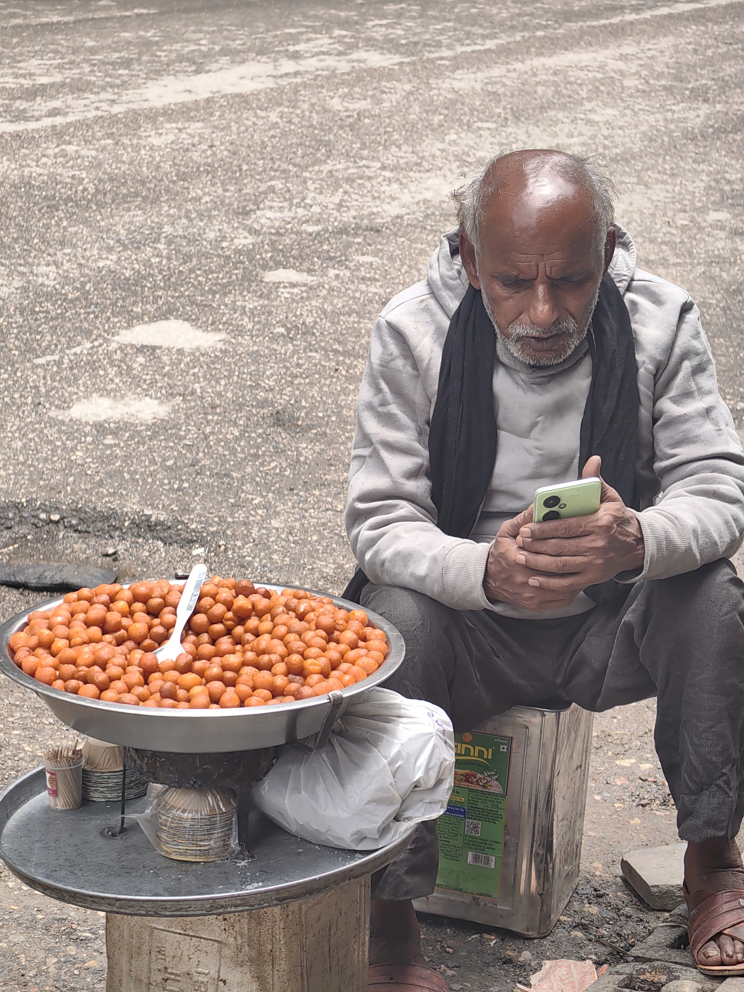 In Manali, India, a street vendor, an elderly man with thinning gray hair, sits on a large metal container. He wears a light grey hoodie and a black scarf. His focus is on his light green smartphone, which he holds in his hands, interacting with it. Beside him, a large metal bowl overflows with numerous small, orange, spherical sweets, likely Gulab Jamun. A white plastic spoon rests atop the sweets. Beneath the bowl, a metal tray holds stacks of small paper cups and a white plastic bag. The scene takes place on a textured asphalt surface, suggesting a street or outdoor marketplace.