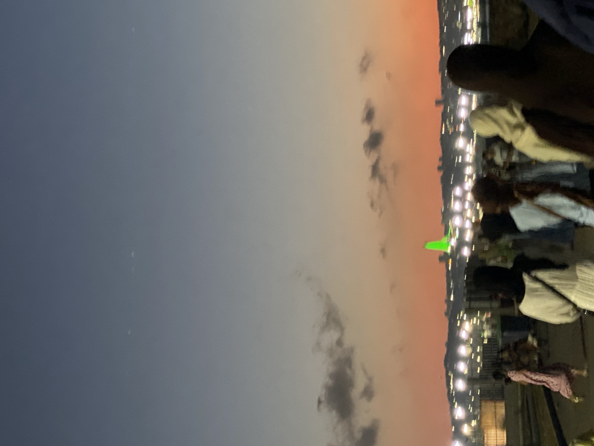 A wide-angle, low-light photograph captures a dusky sky transitioning from deep blue overhead to a vibrant orange horizon. Silhouetted against this twilight are several people walking along a paved walkway. The figures are varied in their attire, with some individuals wearing headscarves. In the background, a city skyline is visible, illuminated by numerous lights. Prominently, a tall, bright green structure, possibly a building or monument, stands out against the cityscape, bathed in a vivid green light. The overall scene suggests an urban evening gathering or stroll, with the city lights and sunset creating a lively yet serene atmosphere. The specific location is identified as Hussein Dey, Algeria.