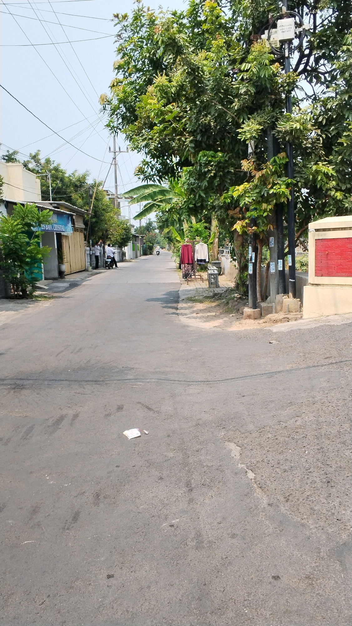 The image depicts a street scene in Banjaragung, Indonesia. A paved road runs through the frame, flanked by buildings and lush vegetation. On the left side, a building with a blue awning that reads "ES BATU CRYSTAL" is visible, with a few people and a motorcycle parked nearby. Further down the street, a distant motorcycle is seen. On the right side of the road, a utility pole with multiple wires and labels stands amidst the trees. Adjacent to the pole, a stand displays two garments, a red skirt or dress and a cream-colored shirt, with a bench placed in front. A wall with a red brick pattern and a yellow border is present at the far right. The sky is clear and blue. A crumpled piece of paper lies on the road in the foreground. The environment suggests a sunny day in a residential or commercial area.