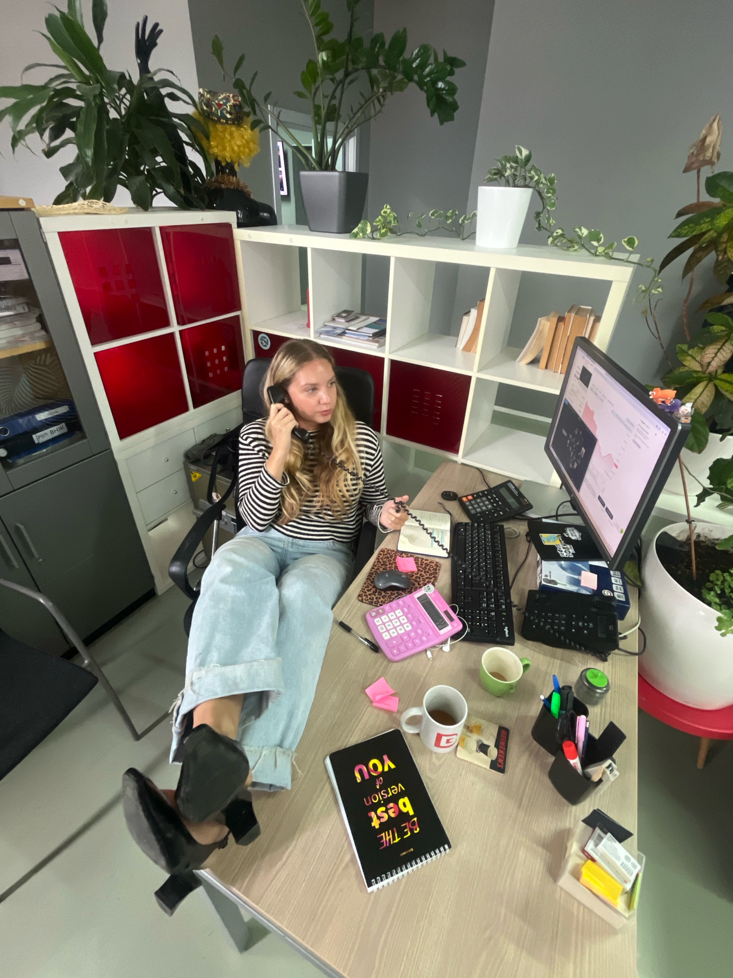 The image shows a woman sitting at a desk in an office environment. She is on the phone, holding a black handset to her ear and speaking. She is wearing a striped long-sleeved shirt and light blue wide-leg jeans. Her feet are crossed, and she is wearing black high-heeled shoes. The desk is cluttered with office supplies, including a computer monitor displaying graphs, a keyboard, a calculator, a pink calculator, notepads, pens, and coffee mugs. In the background, there are shelves with books and decorative items, and several potted plants. The overall impression is of a busy, somewhat unorganized workspace. The location is not specified as Moscow, Russia.