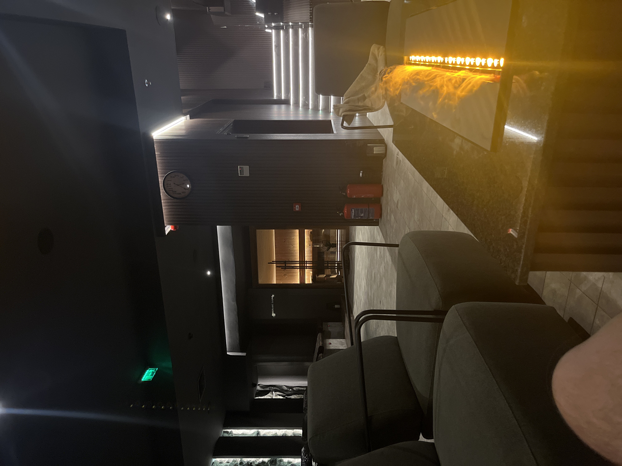 The image depicts the interior of a commercial establishment in Sofia, Bulgaria, featuring lounge seating in the foreground. The seating is upholstered in a dark material and arranged in a row. A stone countertop runs along the right side of the frame, illuminated by an array of linear lighting with a warm hue.
In the midground, a corridor leads deeper into the space, with doors visible along the sides. A fire extinguisher is mounted on the wall near an exit door. An analog clock is also visible on the wall.
In the background, a staircase is illuminated by linear lights, leading to an upper level. The overall atmosphere is dimly lit, creating a sense of privacy and sophistication.