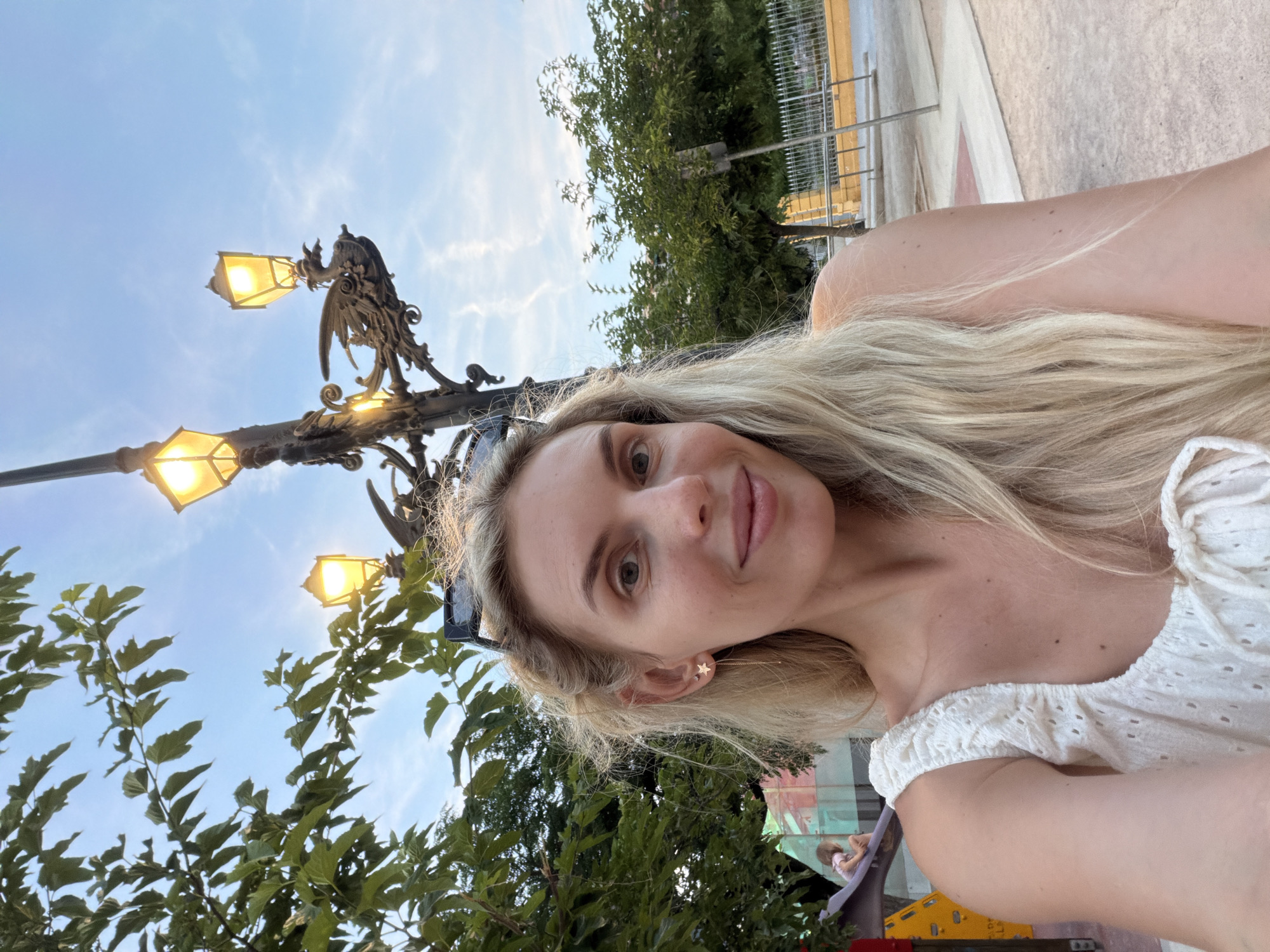 A medium shot captures a blonde woman with fair skin and light blue eyes taking a selfie outdoors. She is smiling slightly and looking directly at the camera. She has blonde hair that falls past her shoulders, and is wearing a white top with eyelet detailing. A gold earring in the shape of a star with a dangling chain is visible in her left ear. Behind her, a streetlamp with three illuminated yellow lights and an ornate, dragon-like fixture is visible against a blue sky with wispy white clouds. To the left, lush green tree branches with large leaves partially obscure the sky. In the background, a playground structure with a red and yellow slide and a young child on the slide can be seen. Further back, trees and a metal fence are present, suggesting an urban park or public space. The woman's location is identified as Benimàmet-beniferri, Spain.