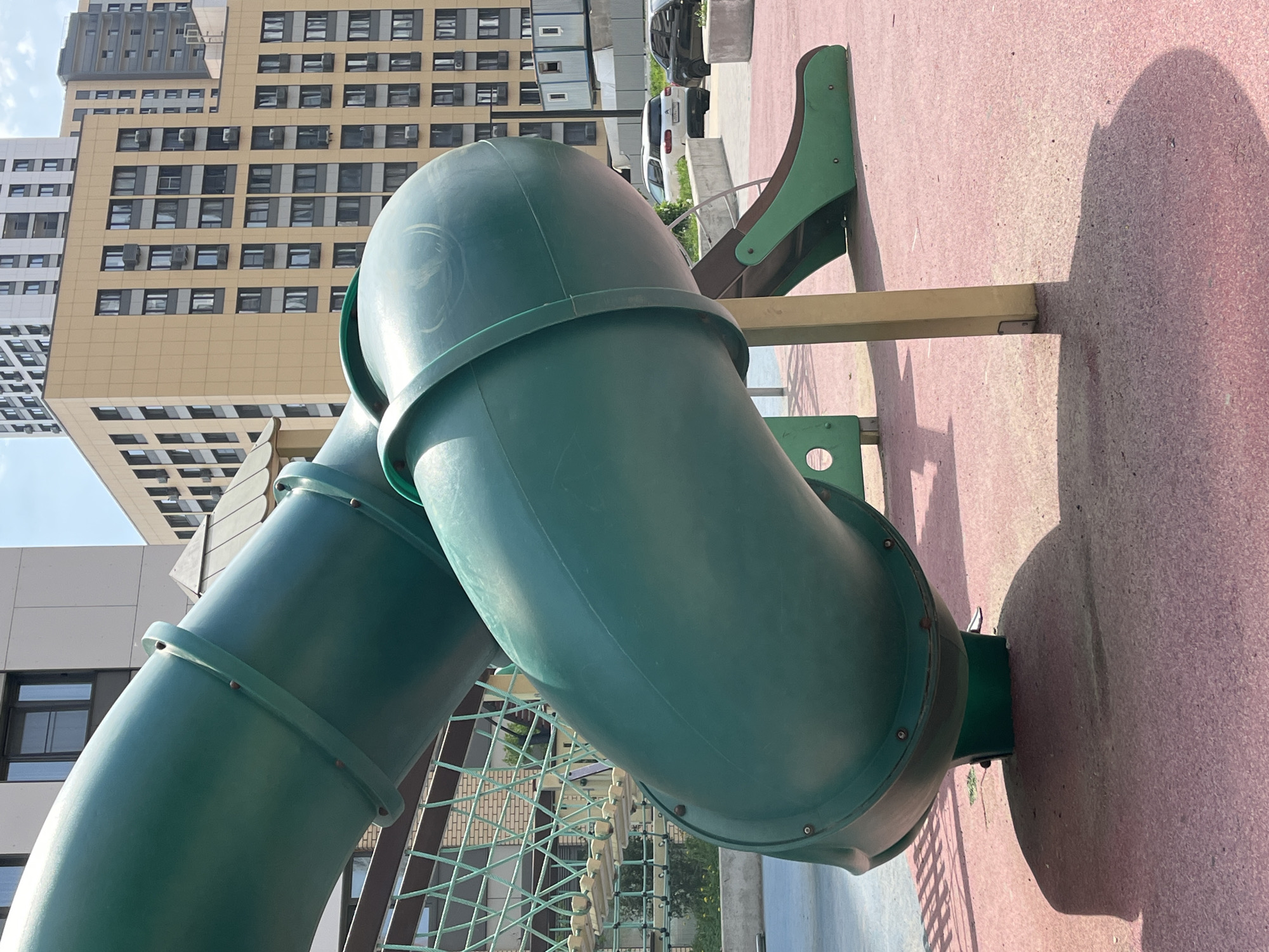 The image depicts a green, cylindrical slide on a playground. The slide is positioned with one end elevated, secured to a platform painted in red. The slide's construction features circular bands along its length, fastened with visible rivets or bolts. A net-like climbing structure is partially visible underneath the slide. In the background, tall buildings are seen, indicating an urban setting. Based on the building architecture and the style of the playground equipment, the location is inferred to be Moscow, Russia. No people are visible in the image. The lighting suggests a sunny day, casting a prominent shadow of the slide on the red platform. Several parked vehicles are on a road in the background, which further confirms a city setting.