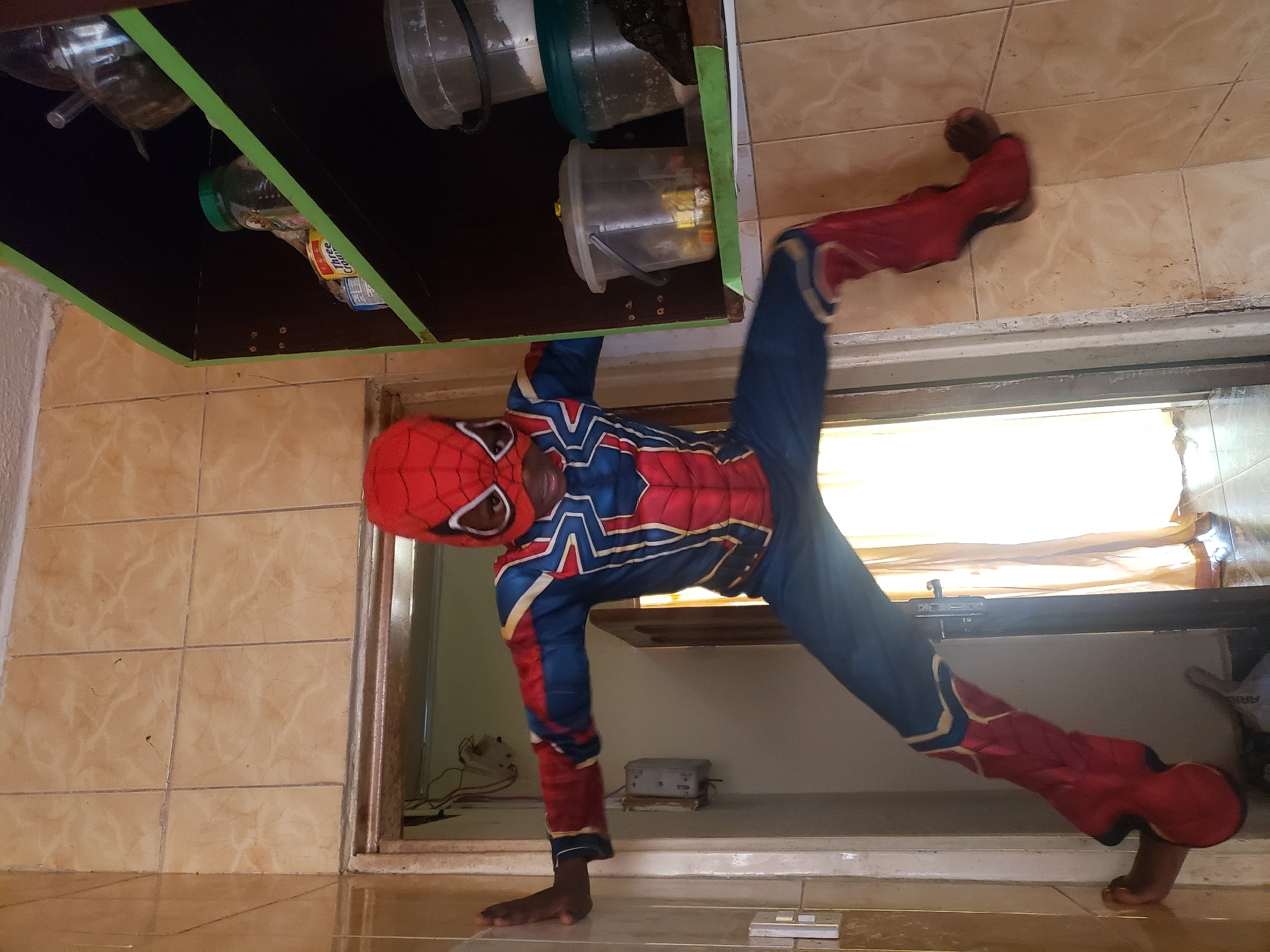 A young Black male, appearing to be between 5-7 years old, is dressed in a full Spider-Man costume and is positioned in a dynamic pose as if clinging to a doorway and a shelf unit. The child's right hand is pressed against a tiled wall to the left of a doorway, and his left arm is extended towards a dark-colored shelf unit. His legs are spread wide, with his right foot braced against the wall to the left of the doorway and his left foot appearing to be supported by the shelf unit or an unseen surface. The costume is red and blue with gold accents and a white spider emblem on the chest. The child's face is visible through the mask, and he is looking towards the camera.
The environment appears to be an indoor space, likely a kitchen or storage area, with beige tiled walls. To the left of the child, a doorway leads to another room, where a yellow curtain is visible. Above the doorway and to the right, a dark shelf unit is filled with various containers and jars. Some items on the shelf include a green-lidded jar, a clear plastic container with items inside, and what appears to be a clear bucket with white contents. Electrical conduits and a white electrical box are visible on the wall next to the doorway. The lighting suggests ambient indoor light, with a brighter light source coming from the doorway on the left. The overall impression is a playful scene of a child in costume, imitating a superhero's actions in a domestic setting. The location is identified as Sabon Tasha, Nigeria.