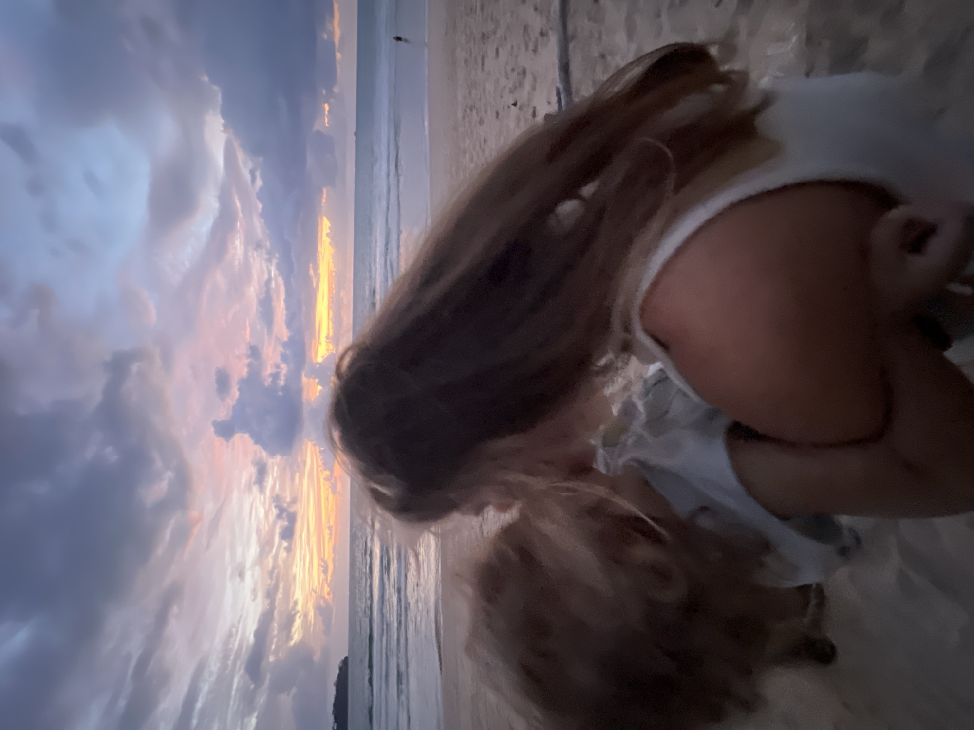 A blurry, close-up shot captures two individuals on a beach at dusk. The person in the foreground, with long brown hair and wearing a white tank top, is embracing another person whose face is obscured by their hair. The background reveals a calm sea meeting a sandy beach, with the sky painted in hues of orange, pink, and purple, indicative of a sunset. The location is identified as Choeng Thale, Thailand.