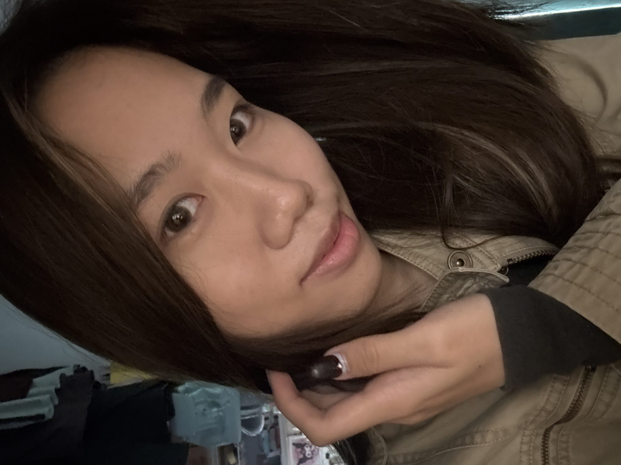 A young woman with long, dark brown hair is positioned in the frame, looking towards the camera. She is wearing a light-colored, possibly khaki, jacket over a dark shirt or sweater. Her hand is near her face, with a painted fingernail visible. The background is partially obscured but includes a glimpse of furniture and what appears to be a window or light source. It is impossible to know the specific location within Nan'an District, China, or to confirm that the image was even taken there, without further information.