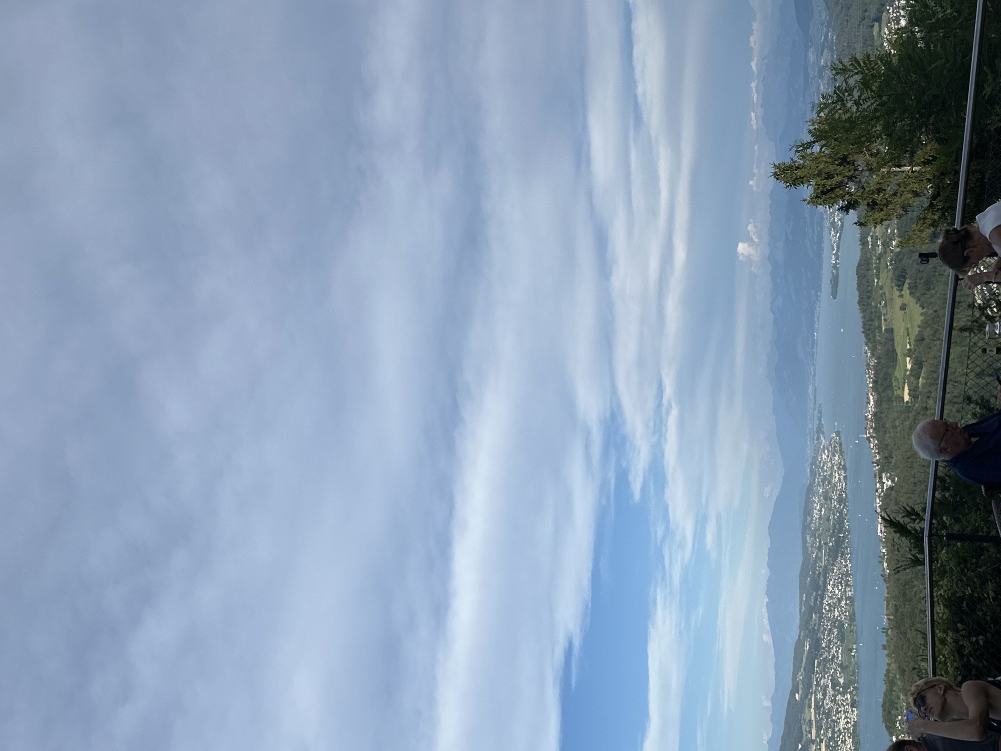 The image captures a panoramic view from a high vantage point overlooking a city nestled along a large body of water, with mountains in the distance. The sky is a mixture of blue and white clouds, with some areas appearing brighter than others. In the foreground, several people are present, seemingly seated or standing near a railing. To the left, a woman with blonde hair and sunglasses is looking upwards, holding a phone in her hand. Next to her, a man in a dark shirt is seated. In the mid-ground, a lush green landscape with scattered trees leads down to the city. The city itself is a densely populated area with many buildings of varying sizes. The water, likely a lake, is a deep blue and appears calm, with a few boats visible on its surface. The distant mountains form a hazy silhouette against the horizon. This scene is characteristic of a view from the area around Stallikon, Switzerland, likely from a restaurant or viewpoint offering vistas of Lake Zurich and the surrounding Alps.
