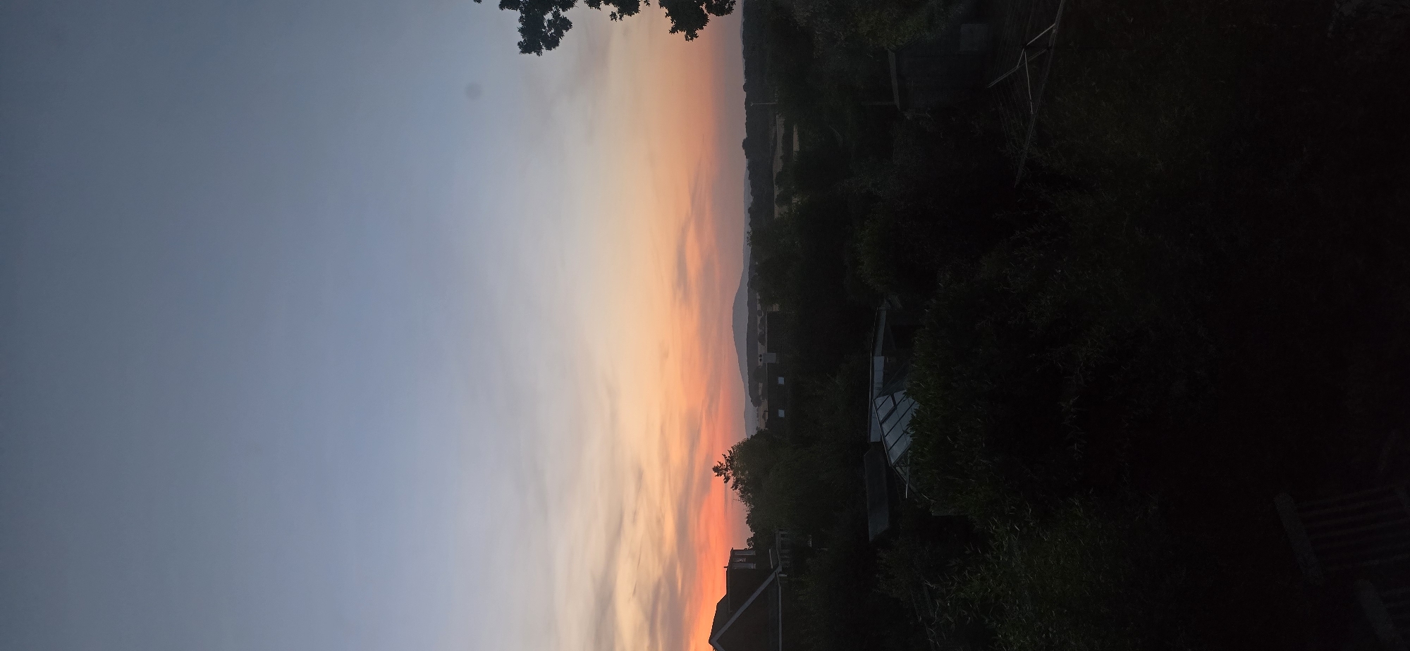 The image captures a sunset scene from Bayston Hill, United Kingdom. The sky displays a gradient of colors, transitioning from a pale blue at the top to hues of orange and pink near the horizon. Silhouetted against the sky are distant hills and trees. In the foreground, a dark mass of foliage obscures much of the lower portion of the image. To the left, the roofline of a building is visible, appearing red-brick. A greenhouse structure is also partially discernible amidst the greenery. The overall impression is one of a rural or suburban landscape at dusk.