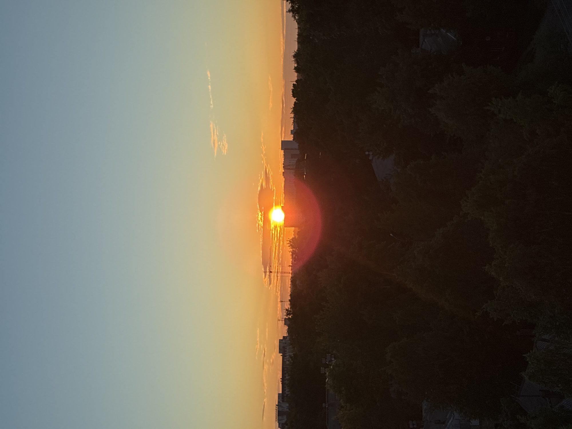 A vertical, eye-level shot captures a sunset over a dense, dark green forest in an urban area. The sun, a bright orange orb, is positioned slightly right of center, partially obscured by thin clouds and emitting a strong lens flare that creates a red halo effect. The sky transitions from a pale blue at the top to a warm orange near the horizon, with wispy white clouds scattered across the transition.
Beyond the forest, a cityscape is visible. Silhouetted against the vibrant sky are several buildings of varying heights and architectural styles. On the right side of the frame, a tall, slender construction crane stands prominently. The foreground is dominated by the dark canopy of trees, with glimpses of rooftops of houses nestled within the foliage. The lighting suggests the golden hour, casting long shadows and intensifying the colors of the sky. The overall atmosphere is tranquil and serene, with a hint of urban life harmonizing with nature.