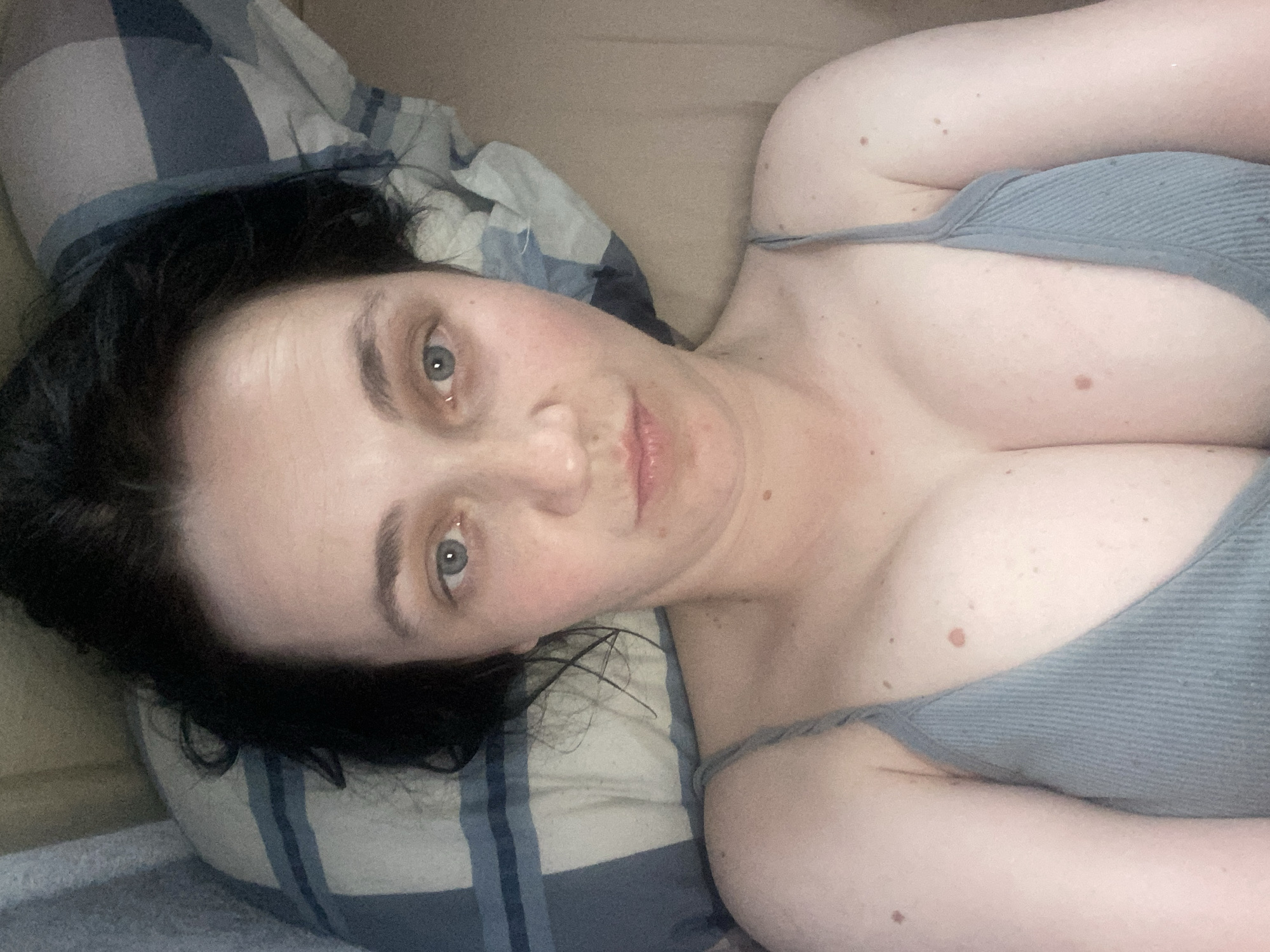 A medium shot, taken from a high angle, captures a woman lying down with her head on a pillow. She has dark hair and blue eyes. Her face is centered in the frame, and she is looking directly at the camera. She is wearing a light gray ribbed tank top with thin straps. The background consists of a pillow with a blue and white plaid pattern and a plain brown surface. The lighting is soft and diffused.