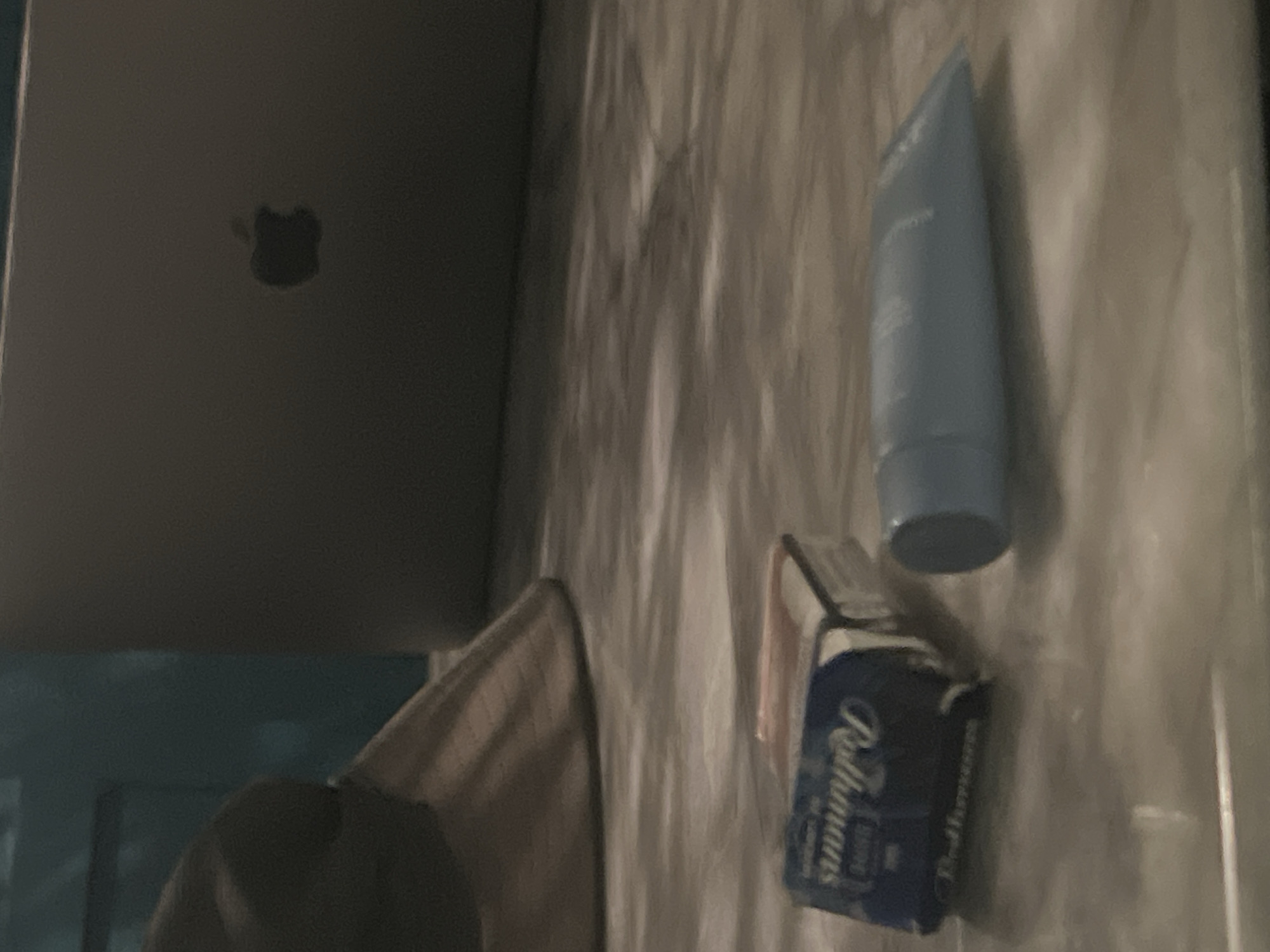 The image displays an indoor scene, likely a flat lay, featuring several objects on a textured surface. The overall lighting is dim.
On the left side of the frame, occupying a significant portion, is the dark gray back of a laptop, identifiable by a prominent, dark Apple logo situated centrally.
On the right, two items are visible. Towards the top right is a light blue, cylindrical tube, typical of a cosmetic or personal care product. Faint text on its surface suggests the brand "DOVE", possibly indicating "moisturizing cream" or similar. Below and slightly to the left of the tube is a small, open cardboard box. This box is predominantly dark blue with white accents. Visible text on the box clearly reads "Rothmans" in a stylized script, and below it, "KING SIZE" and "MENTHOL", indicating it is a cigarette packet. The top flap of the box is open, revealing a white interior.
The objects rest on a light-colored, patterned surface, which appears to be a marbled countertop or similar table surface. In the bottom left corner, part of a light-colored fabric or towel is visible.
There is no activity or event taking place; the items are stationary. The time of day cannot be definitively determined, but the dim lighting suggests it could be indoors with artificial light or low natural light. No weather conditions or emotions are discernible. While the location context is given as Abuja, Nigeria, there are no visual cues within the image itself to confirm this.