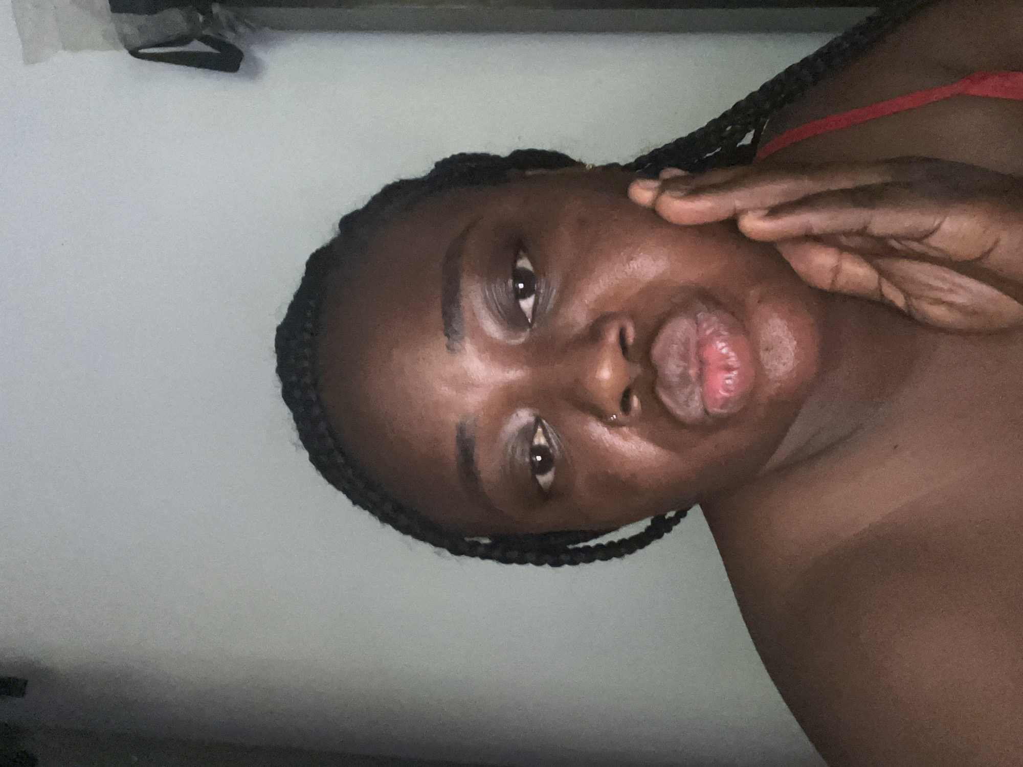 A close-up, portrait-style photograph, rotated 90 degrees clockwise, features a dark-skinned individual, likely female, in an indoor setting. The subject has braided hair and is looking directly into the camera with a neutral to slightly pensive expression. A silver nose piercing adorns her left nostril. Her full lips are slightly parted, revealing some texture. One hand, with visible fingers, gently rests on the right side of her face near the cheekbone. A red strap, possibly from a top or undergarment, is visible on her shoulder. The background is a plain, light-colored wall, with an indistinct dark object in the top left corner. The lighting is soft and somewhat dim, casting subtle shadows. No text is visible. The provided context indicates the location is Abuja, Nigeria.