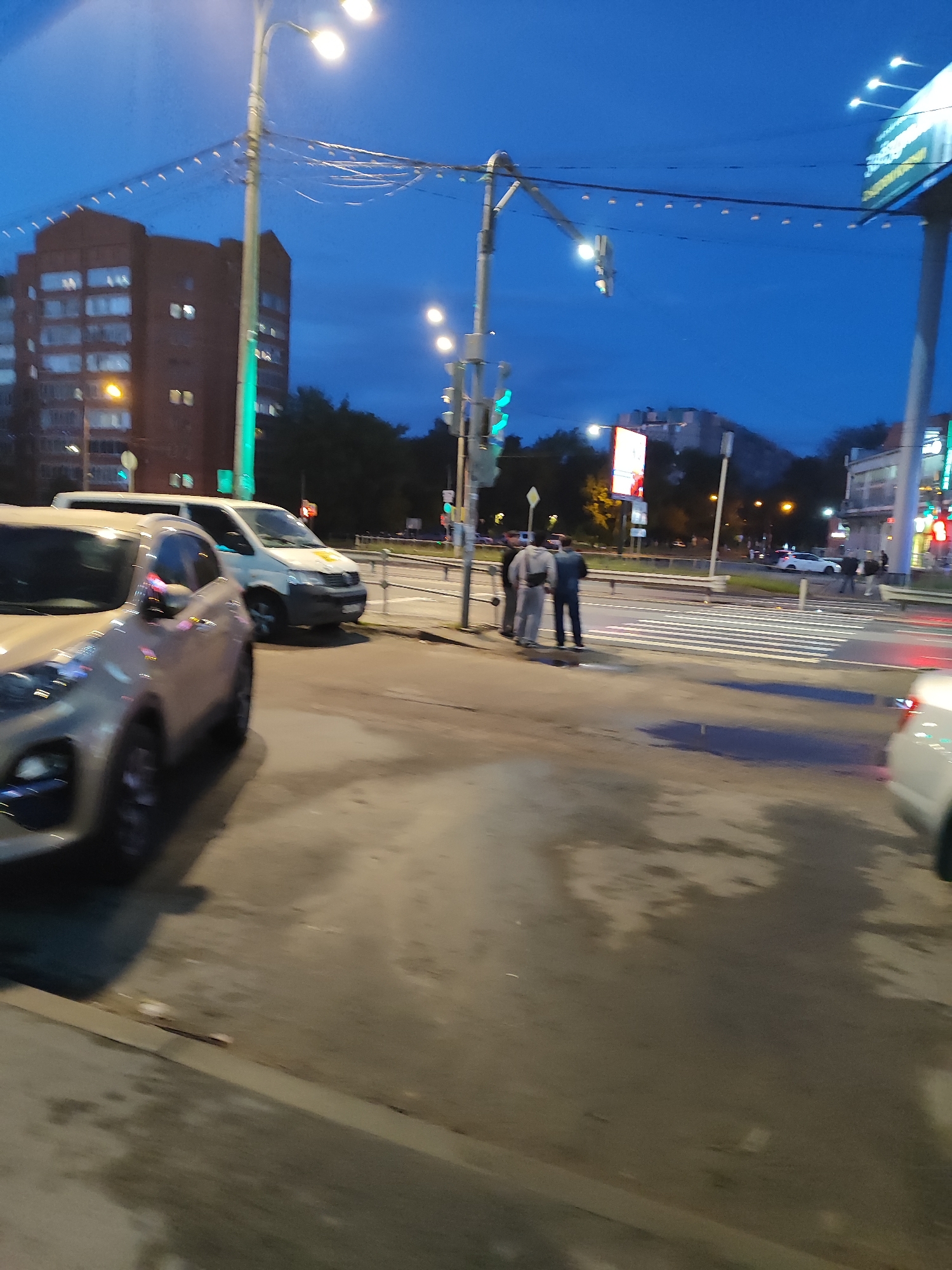 The image depicts an urban street scene at dusk or night. In the foreground, a light-colored car is partially visible on the left, with its headlights reflecting on the wet asphalt. A white van is parked behind it. In the middle ground, a pedestrian crossing is marked with white stripes. Several individuals, dressed in casual clothing, are standing near the crosswalk. One group of three men is clustered together, while other pedestrians are further away. In the background, buildings of varying heights are illuminated, and streetlights cast a glow on the scene. A large billboard with colorful graphics is visible on the right side of the image. The sky is a deep blue, indicating twilight or night. The ground appears wet and reflective, suggesting recent rain. The overall atmosphere is that of a typical urban intersection at the end of the day.