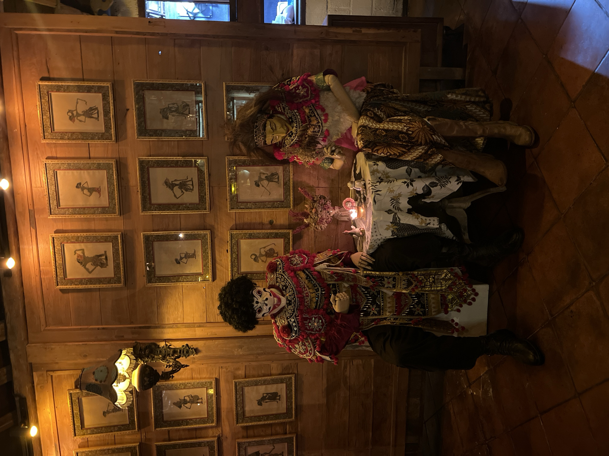 A medium indoor shot shows two mannequins, one with a long brown wig, lying prone on a table with its head to the left, and the other with a black afro wig, also lying on the table, head to the left, in a North Kuta, Indonesia establishment.
The background consists of a paneled wall covered with framed monochrome art. A hanging light fixture is visible to the lower-left. The tile floor is visible on the lower-right.