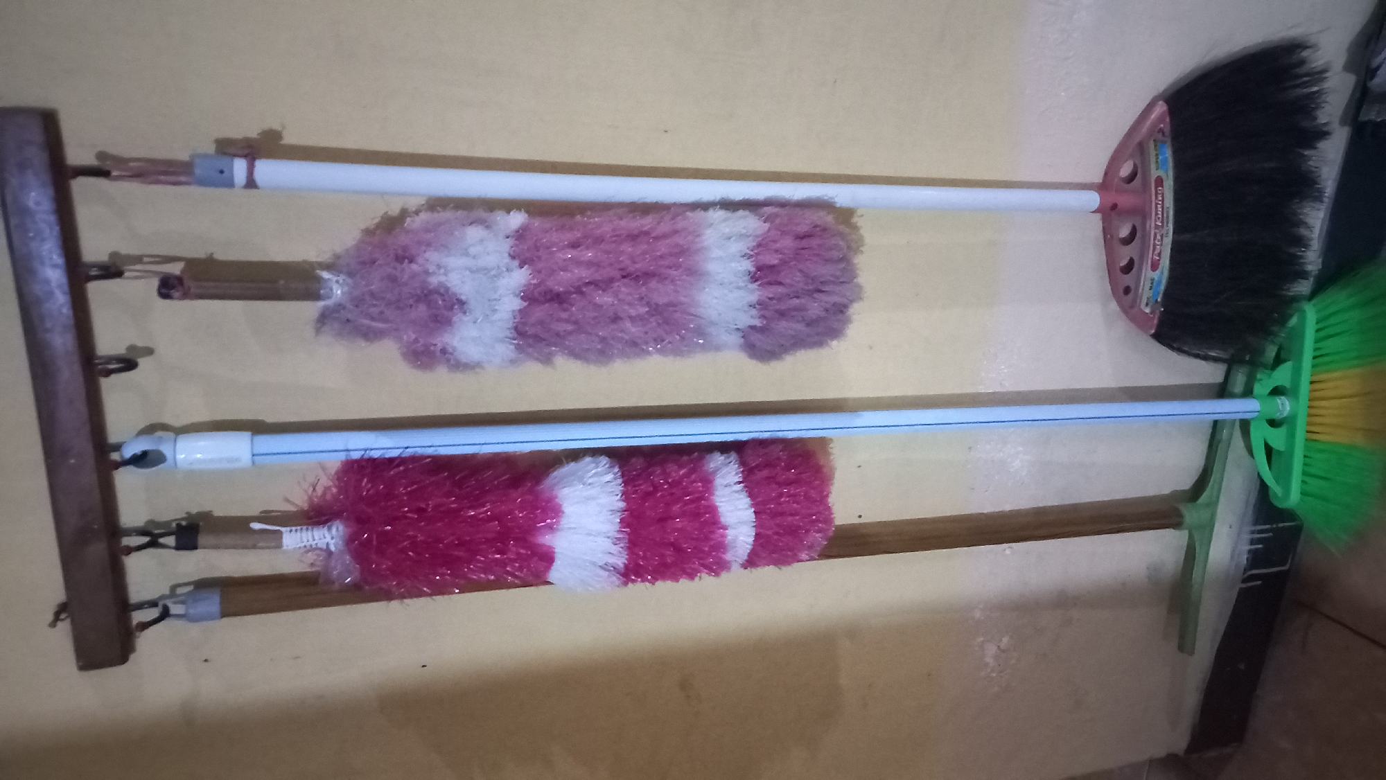 The image depicts a collection of cleaning tools, including several brooms and dusters, arranged in a vertical orientation against a textured, pale yellow wall. A wooden rack with multiple hooks is mounted horizontally near the top of the frame, from which the items are suspended.
On the left, a broom with a wooden handle and green bristles is partially visible, resting on the floor. Above it, a pink and white striped duster hangs from a hook. To its right, another duster, also pink and white striped, is suspended. Beside this, a broom with a blue handle and green and yellow bristles stands upright, its head touching the floor. To the far right, a broom with a white handle and black bristles is also standing upright. The floor appears to be a tiled or concrete surface.
The scene suggests a storage area for cleaning supplies, possibly in a home or a small establishment. The lighting is somewhat dim, creating shadows on the wall. No people are present in the image. The image context does not provide any information about the specific location in the city of Pare, Indonesia.