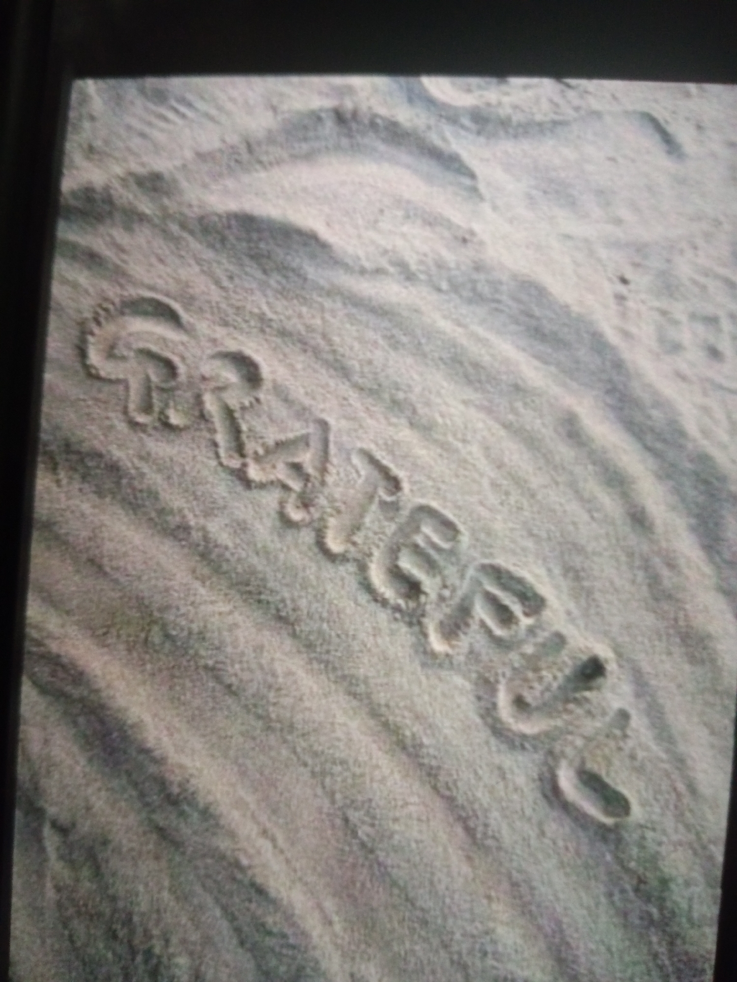 The image depicts the word "GRATEFUL" written in the sand. The letters are formed by indentations in the sandy surface, which appears textured with ripples. The lighting suggests it may be daytime. The image is a close-up, focused on the text in the sand. There are no people visible in the frame. The environment is a sandy area, possibly a beach or a desert, with subtle shadow patterns indicating the contours of the sand. There is no information to suggest this location is in Jalingo, Nigeria.