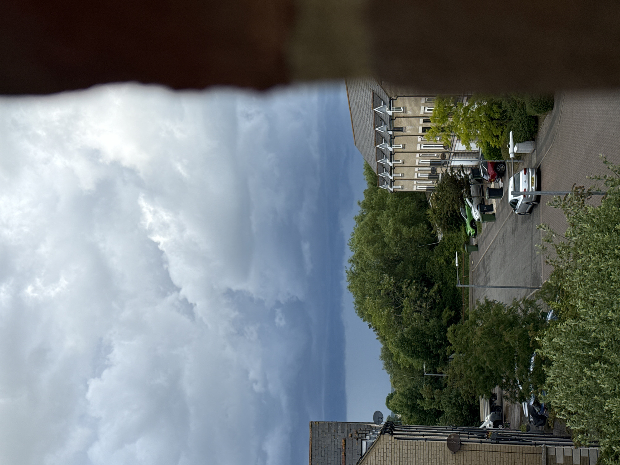 This media file presents a residential street scene, viewed from an elevated position, possibly a window or balcony, with a dark, blurry obstruction across the top, framing the shot. The scene depicts a predominantly overcast sky, with a dramatic mix of bright white and darker grey clouds suggesting impending or recent rain. Some areas of blue sky are visible through breaks in the cloud cover.
To the right, a row of multi-story residential buildings with light-colored brick or rendered facades and dark, sloping tiled roofs are visible. Many windows with white frames line the buildings, and some feature small, decorative white triangular elements beneath the roofline. A satellite dish is affixed to one of the roofs.
Below and to the left of the buildings, a paved street, constructed from interlocking brick pavers, winds through the area. Several cars are parked along the street. A silver hatchback, possibly a VW Polo or Golf, is prominent towards the center-right, displaying a partially visible yellow UK license plate beginning with "YJ6" and what appears to be a disabled parking badge on its dashboard. Further left, a green car, possibly a small SUV, and a dark red car are also parked. A green waste bin is situated near the green car.
The scene is rich with greenery, featuring numerous mature trees with dense, varied foliage bordering the street and interspersed among the buildings. Utility poles or lamp posts are also visible along the street.
No people are visible in the image. The lighting suggests daytime, likely an overcast afternoon, consistent with the location context of Great Yarmouth, United Kingdom. The overall atmosphere is quiet and typical of a suburban residential area.