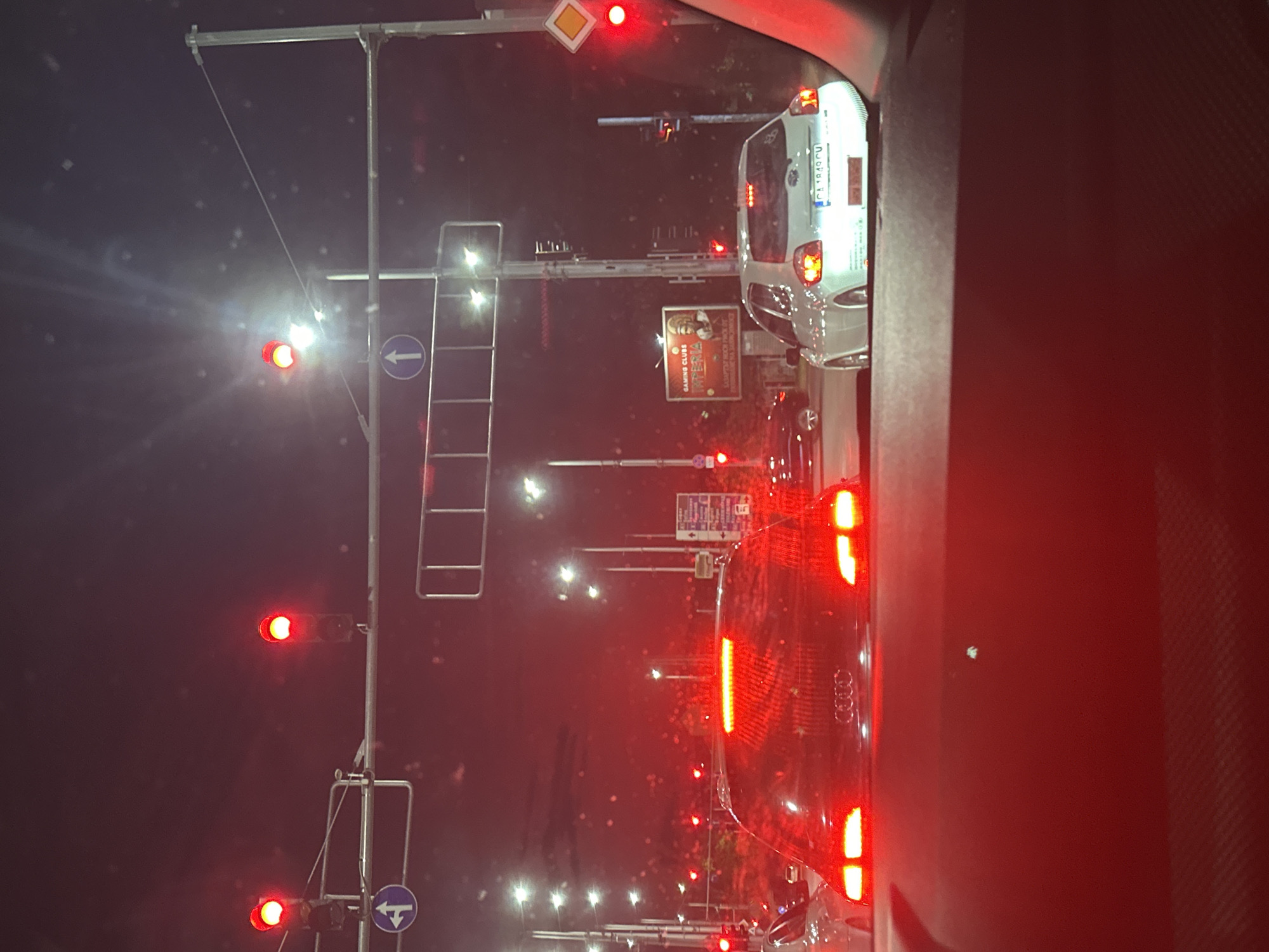 The image shows a traffic scene at night in Plovdiv, Bulgaria. The perspective is from inside a vehicle, looking forward through the windshield. Traffic lights are red for all visible lanes, indicating a stop. Several cars are stopped in front, including a black Audi with its brake lights illuminated and a silver hatchback further ahead with its license plate partially visible. Streetlights and the illuminated sign for "GAMING CLUBS INTERNA" are visible in the background. The overall atmosphere is dark with strong red light from the traffic signals and vehicle lights. The location is likely an intersection on a main road in Plovdiv, given the presence of traffic lights, multiple lanes, and signage.
