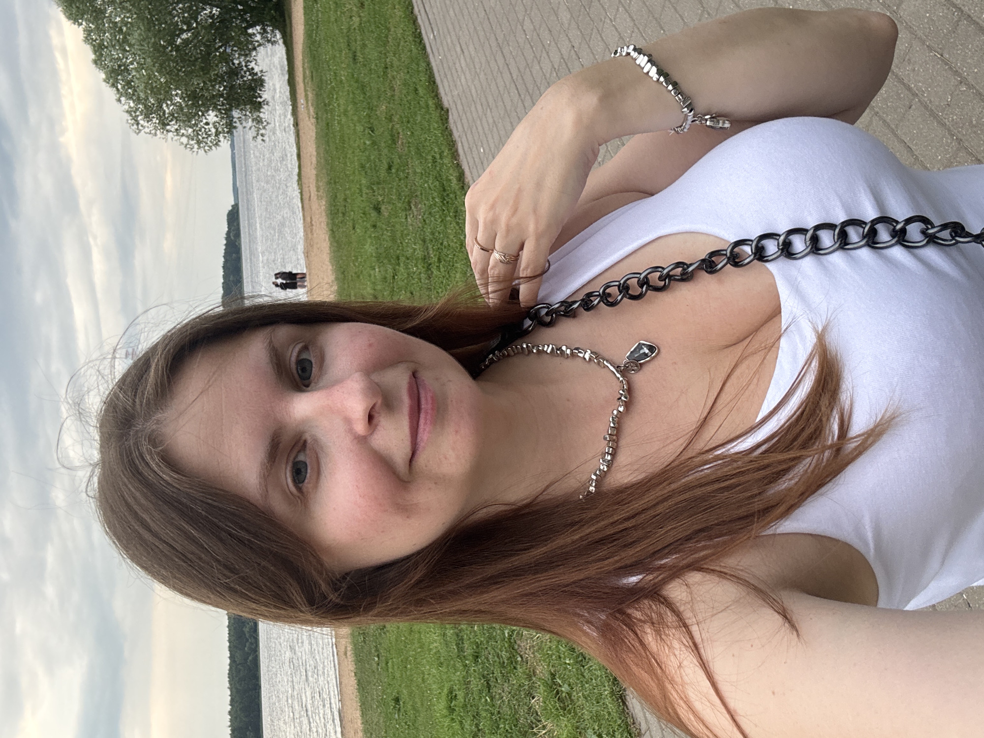 A medium shot, outdoor selfie shows a young woman in a white tank top standing on a paved walkway near a body of water, likely a river or lake. She has shoulder-length brown hair and is wearing a silver necklace and bracelet. She is smiling and looking at the camera, with her right hand resting on her neck. In the background, there is a sandy beach, a grassy area, and trees. The sky is overcast. A couple of people are visible in the distance on the beach. The setting is likely a park or recreational area in Konakovo, Russia, given the presence of water and trees typical of the region.