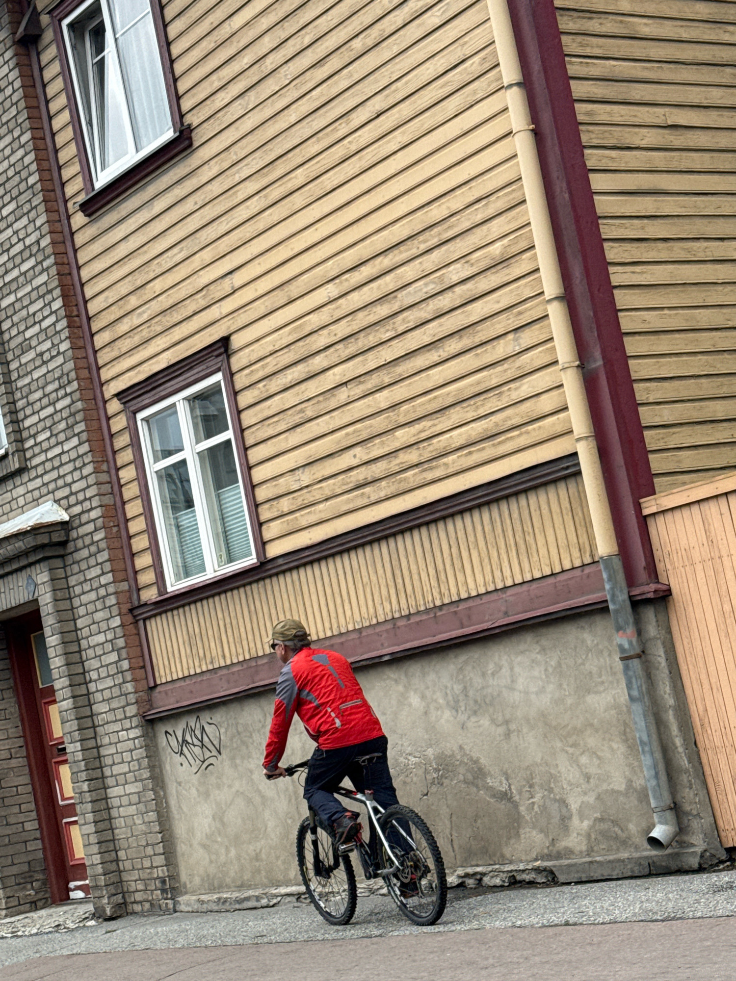 A man wearing a red jacket, blue jeans, and a baseball cap rides a bicycle past a weathered, tan wooden building with a dark red trim. The building has several windows with white frames and blinds. To the left of the wooden building is a brick building with a dark red door. The ground is paved with grey and white cobblestones. There is graffiti on the concrete wall behind the man. The scene appears to be in an urban setting in Tallinn, Estonia, given the architectural style.