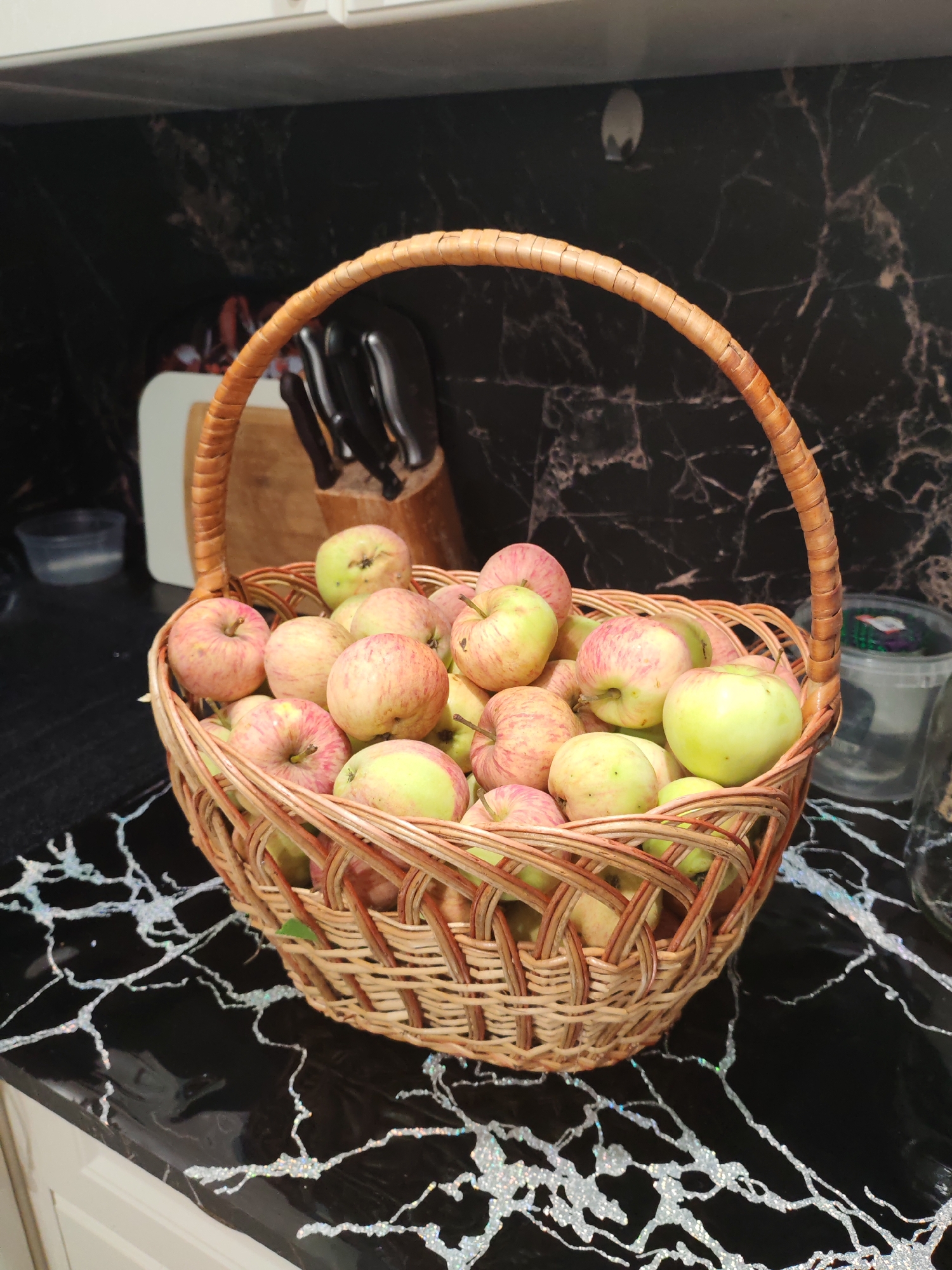 A wicker basket filled with ripe apples sits on a black marble countertop. The apples are a mix of green and red, some with pink blush. The background shows a kitchen scene with a knife block and various containers. The countertop has a distinctive white, marbled pattern. The lighting is natural and appears to be from an overhead source. The overall impression is one of abundance and harvest.