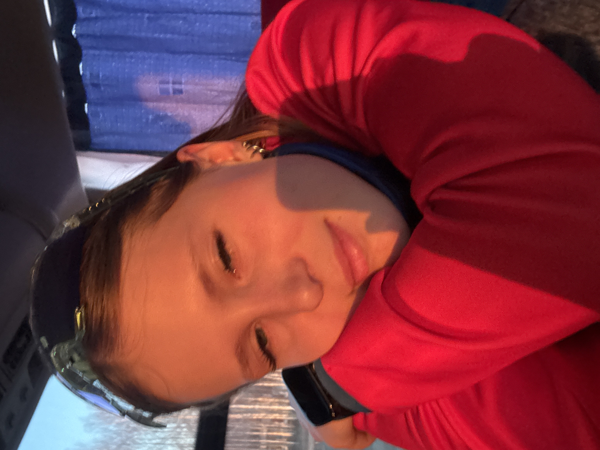A young person, likely a female, is sleeping in a vehicle. She is wearing a red hooded sweatshirt and has sunglasses pushed up on her forehead. The background shows what appears to be a blue curtain or blind, possibly inside a bus or train. There is no clear evidence to confirm a location in the city of Osa, Russia from this image alone.