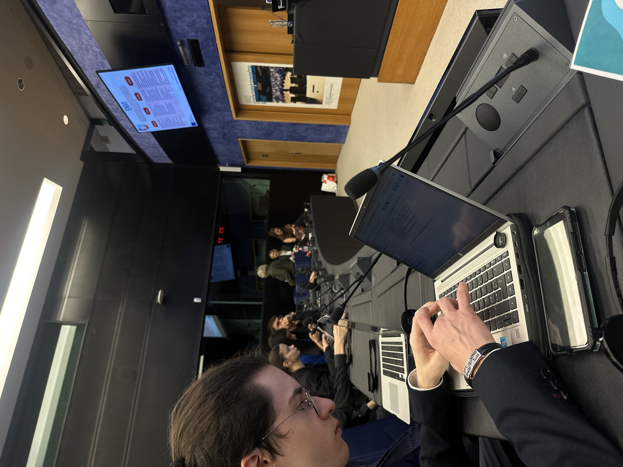 The image depicts a meeting in Strasbourg, France, likely at the European Parliament based on the location and signage. A row of individuals, some wearing headsets, are seated at desks equipped with laptops and microphones. One individual in the foreground is focused on typing. A monitor displays the word "ENVI," suggesting a committee or topic related to the environment. The room's decor includes wood paneling, a poster, and a blue patterned wall.