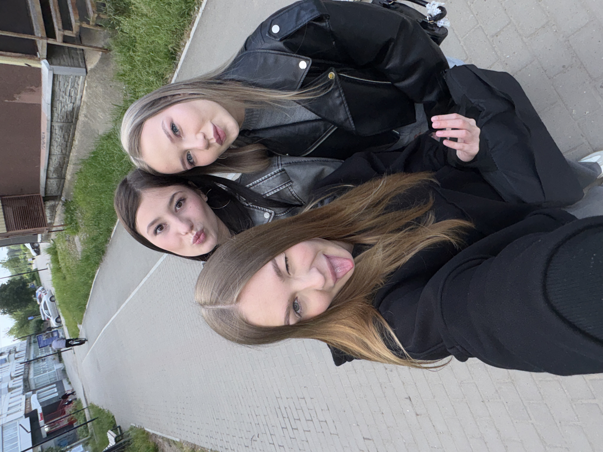 Three young women are posing for a selfie outdoors. The woman in the foreground is sticking her tongue out and winking. Behind her, another woman is pouting her lips. The third woman is partially obscured. All three women are wearing dark clothing, including jackets. The background includes a paved walkway, grass, buildings, and parked cars. The buildings appear to be of modern construction. Based on the details in the image, this appears to be in Chaikovsky, Russia.