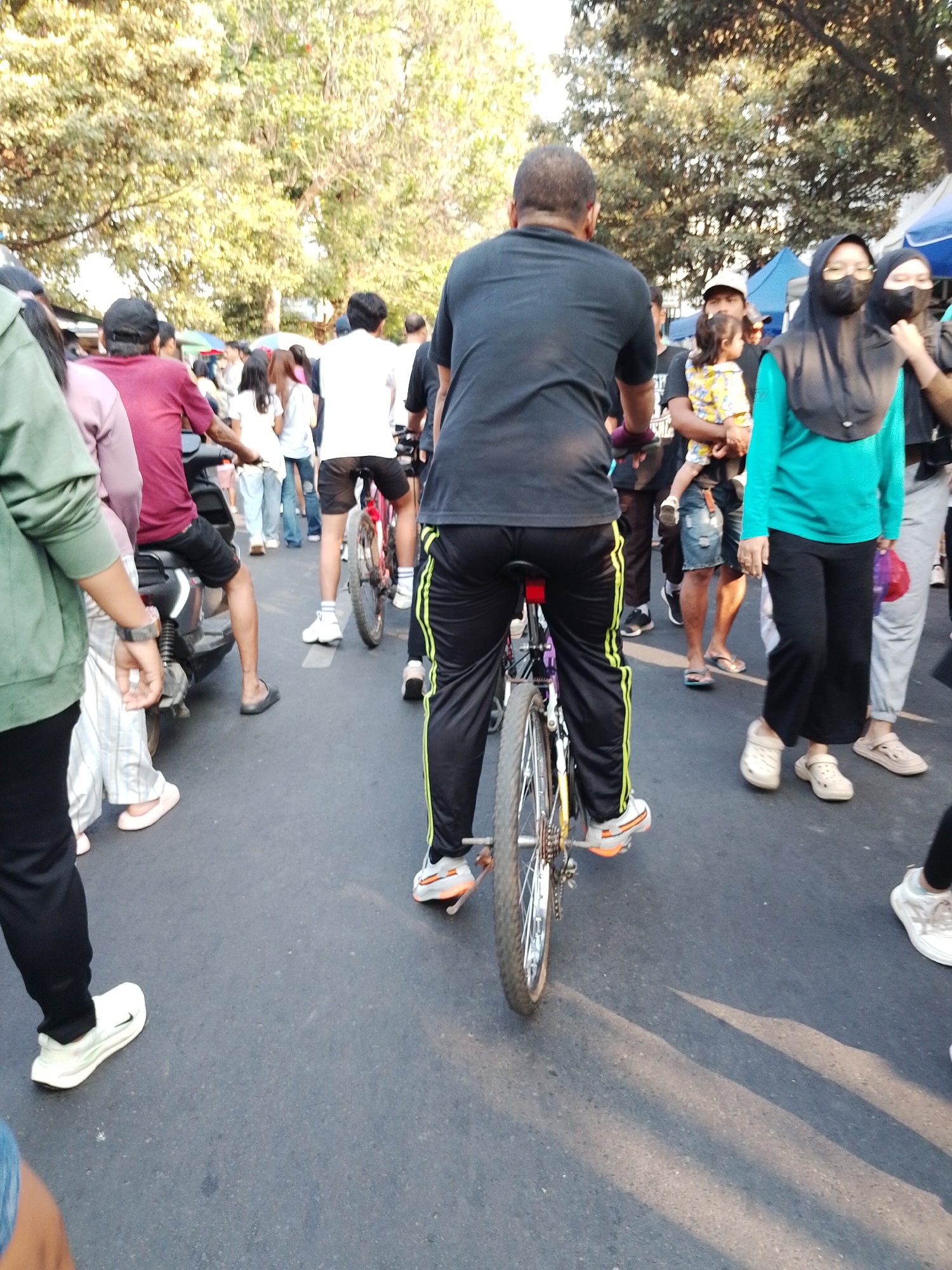 This is a medium shot, eye-level, outdoor photograph taken during the day in Kecamatan Pati, Indonesia. The scene depicts a street with people and bicycles. In the foreground, a man wearing a black t-shirt and black pants with lime green stripes is riding a bicycle. The bicycle has a yellow frame and a silver rear wheel. To the left of the bicyclist, a person wearing a green hoodie and black pants stands facing away from the camera, their left side visible. Further to the left, a person in a maroon t-shirt and black shorts is seated on a motorcycle, also facing away. In the background, a crowd of people are walking and standing. Some individuals are wearing face masks, including a woman in a teal top and black hijab. Several trees with dense green foliage are visible overhead, with sunlight filtering through. The ground is paved asphalt, and shadows from the trees and people are cast across it. The overall atmosphere appears to be that of a busy street or event.