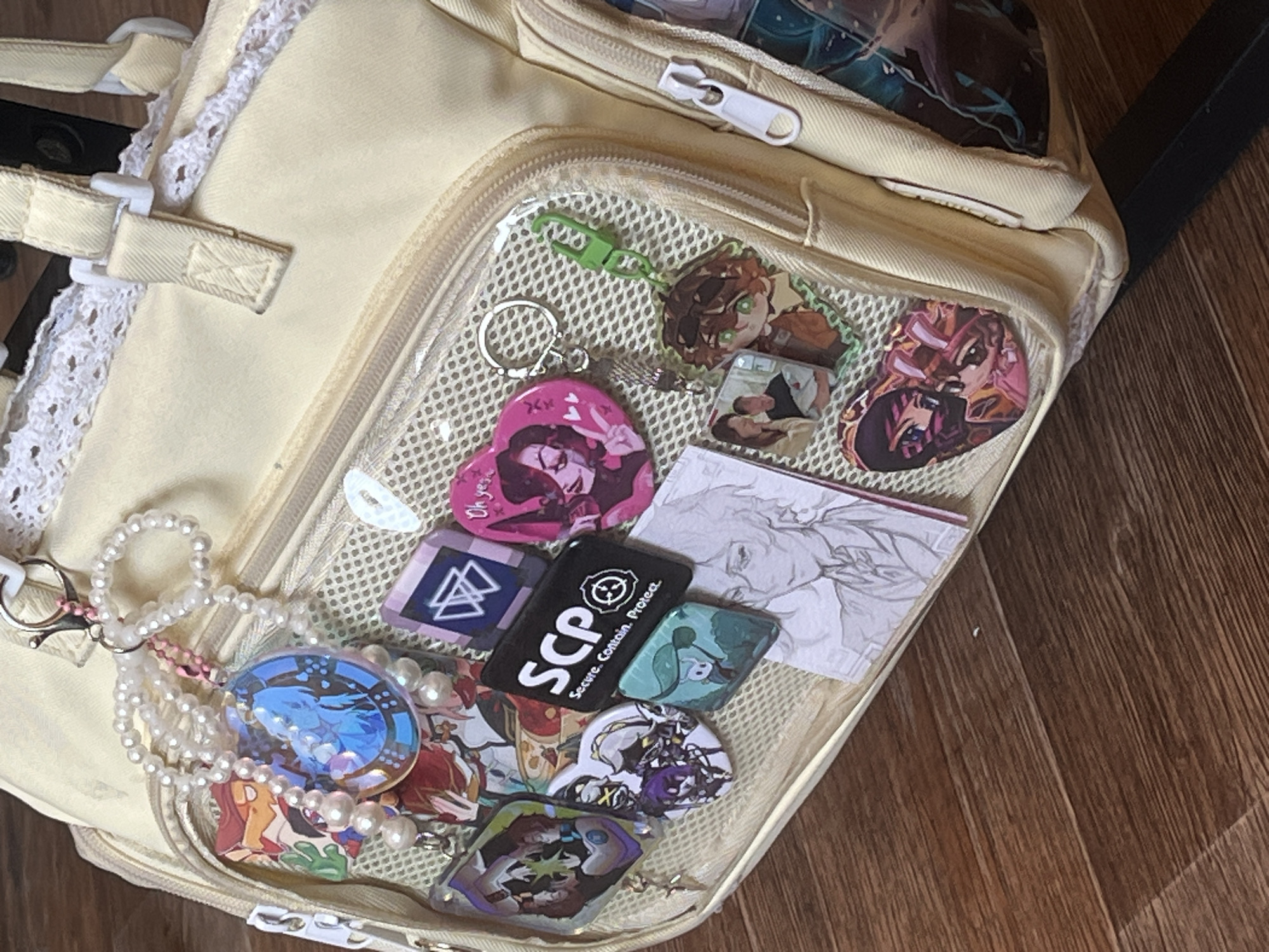 A close-up shot depicts a cream-colored backpack placed on a brown wooden surface. The backpack features a transparent front pocket filled with various items, including keychains, pins, and paper items. The keychains and pins display a variety of designs, with some depicting anime characters or logos. One prominent item is a black pin with the letters "SCP" in white, along with the phrase "Secure. Contain. Protect." There is also a small string of white pearls and other colorful beads attached to the backpack. The backpack has white lace trimming on the left side.