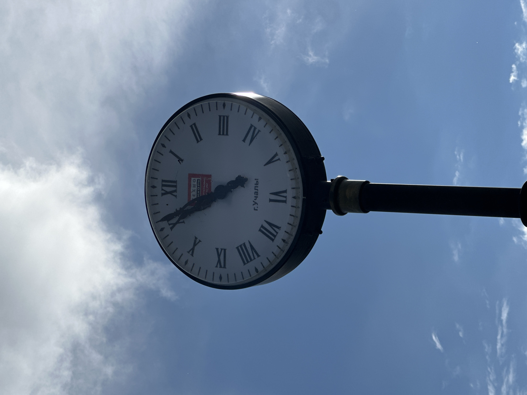 A large, round clock with Roman numerals dominates the center-left of the image, seen from below against a bright, partially cloudy sky. The clock face is white with black Roman numerals marking the hours from I to XII. The short hour hand points just before the IX, while the long minute hand points directly at the X, indicating the time is approximately 8:50.
Visible on the clock face is Cyrillic text. At the top-center, "Г. УЧАЛЫ" is clearly legible, identifying the city as Uchaly. Below this, within a red rectangular emblem, more Cyrillic text can be seen. The first line appears to be "ТЕХНО", followed by text resembling "ИЮЛЬ" or "ИЗОЛ", and then "СТРОИТЕЛЬНЫЕ" (meaning "building") and "МАТЕРИАЛЫ" (meaning "materials"). This suggests a company or sponsor related to building materials, possibly "Technoizol Building Materials".
The clock is mounted on a dark, cylindrical pole extending from the right side of the frame. The sky transitions from a clear blue on the right to an expanse of white, wispy clouds on the left. Bright sunlight reflects off the upper rim of the clock, suggesting a sunny daytime environment. There are no people visible, and no specific event is taking place, just the static display of the time. The overall impression is one of an urban or public setting on a bright day.