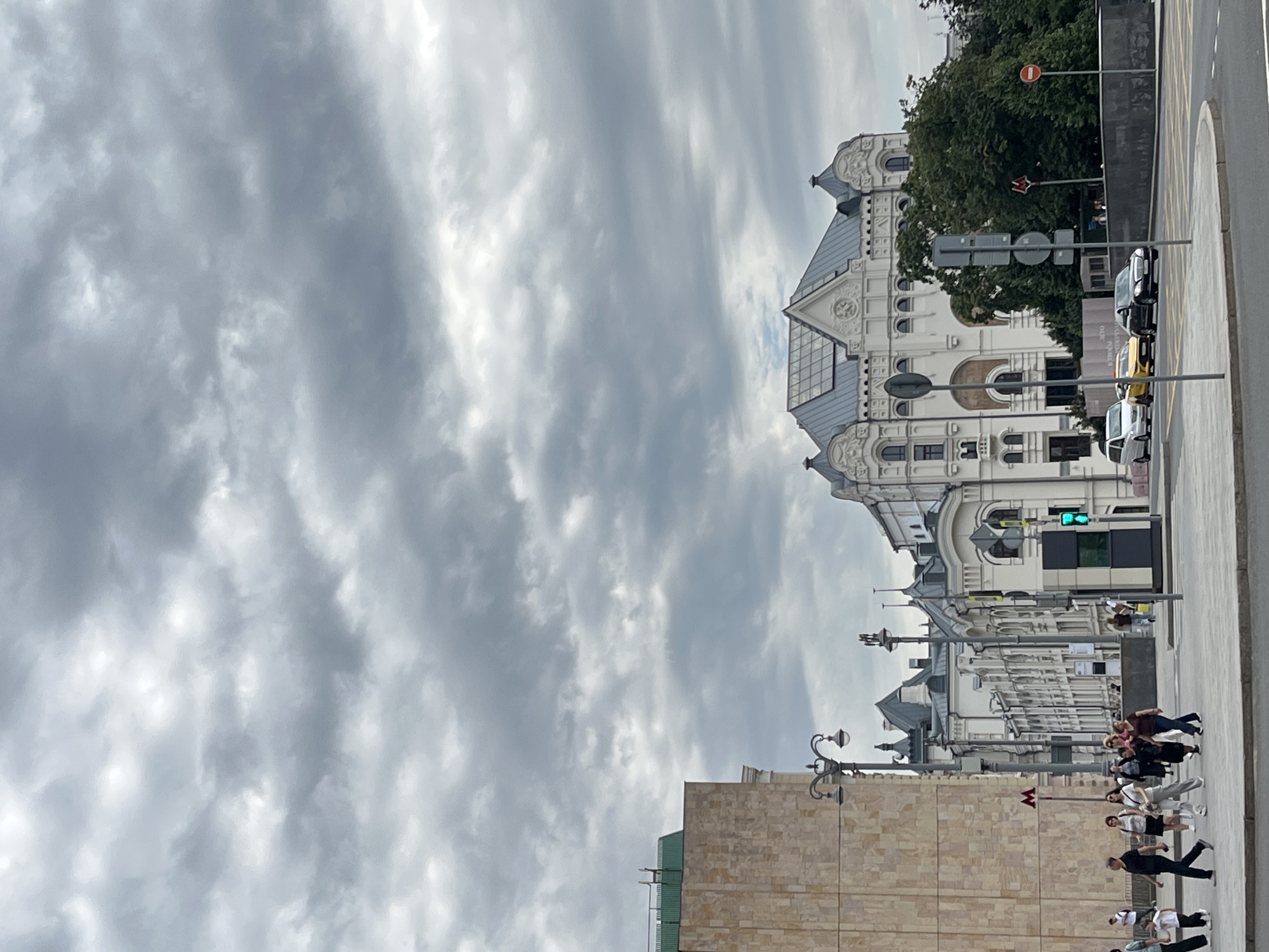 The image captures a street scene in Moscow, Russia, under a heavily overcast sky filled with dramatic grey clouds. In the foreground, a paved pedestrian walkway runs parallel to a street where several cars are visible, including a white van, a yellow taxi, and a black sedan. Pedestrians are crossing the street at a marked crosswalk.
Dominating the background is an ornate white building with a multi-gabled roof, featuring intricate architectural details, arched windows, and decorative facades. To the left of this building, a large, modern structure with a beige, textured exterior rises, topped with a green roof.
Street furniture includes lampposts with ornate fixtures, traffic lights displaying a green signal, and various street signs. Trees line the background, some of which appear to be quite large and leafy, contrasting with the urban architecture. A red metro sign is visible on a pole, indicating proximity to a subway station. The overall impression is that of a bustling urban environment under a moody, cloudy sky.