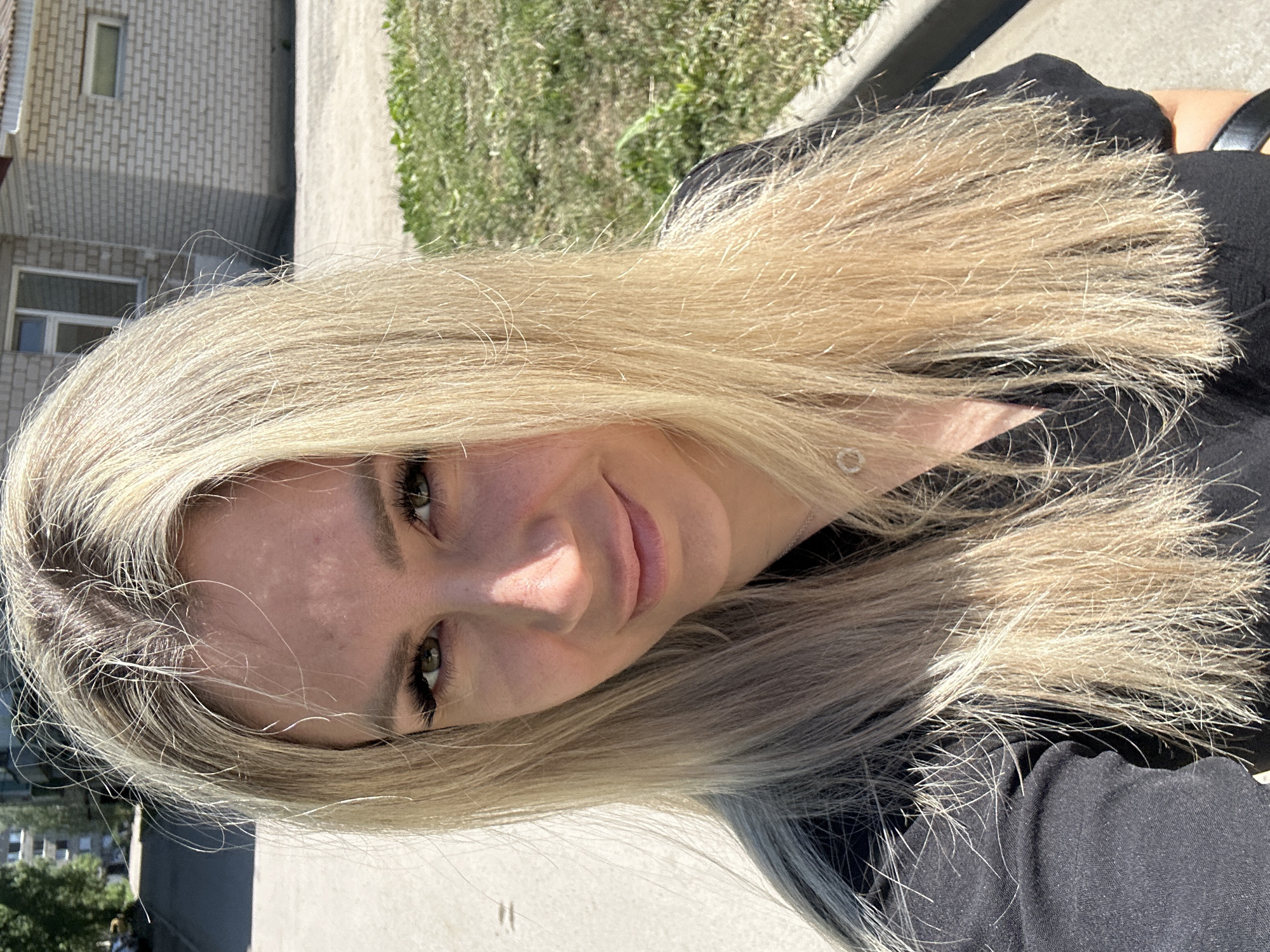 A medium shot selfie is taken outdoors during daylight. The subject is a blonde woman with long hair, wearing a black top. She is looking at the camera with a slight smile. Her face is illuminated by the sun, casting a shadow on the left side of her face. The background shows a paved area, a grassy area, a curb, and a brick building with windows. In the distance, trees and buildings are visible, suggesting an urban or suburban environment. The overall scene is bright and clear.